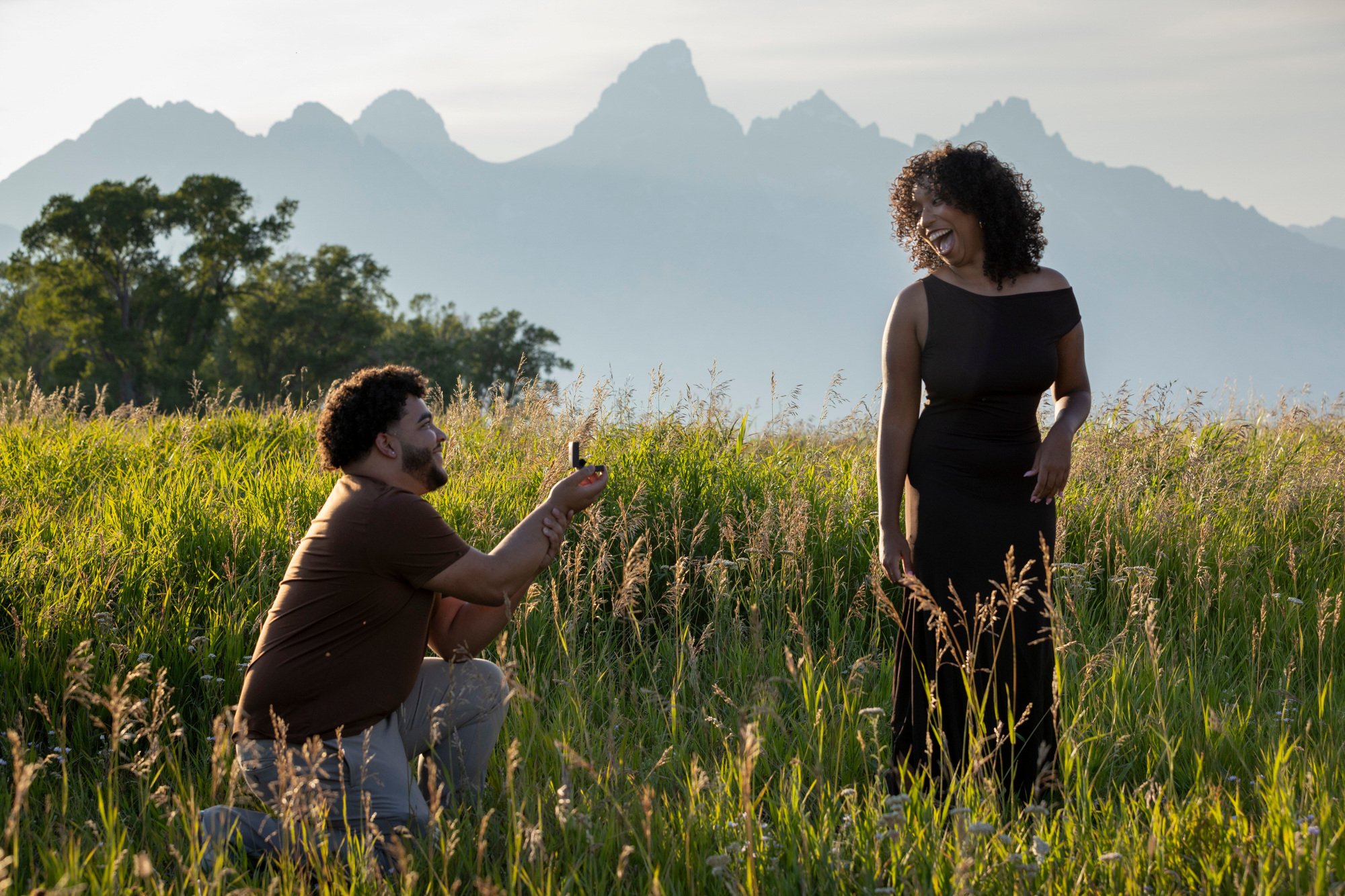 A woman in a black dress laughing as a man kneels on grass taking her photo with a smartphone, with mountains in the background.