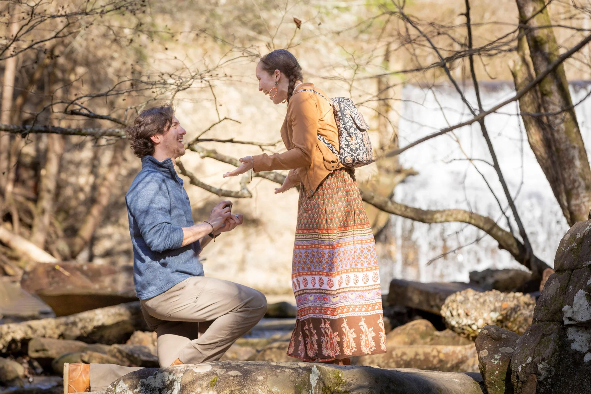 A man is kneeling on one knee in front of a woman, proposing marriage on a rocky creek bed in a forest with autumn leaves, while both are smiling and facing each other.