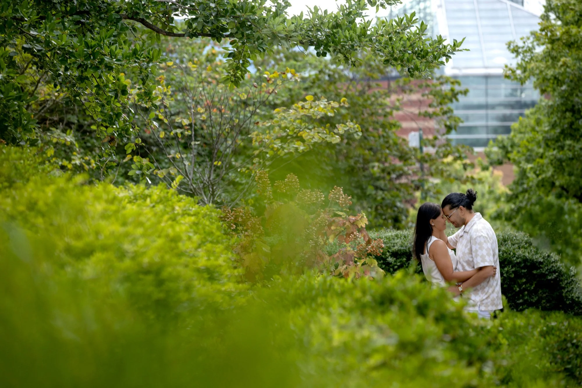 A couple is standing close together amongst green bushes and trees in an outdoor park, with city buildings visible in the background.