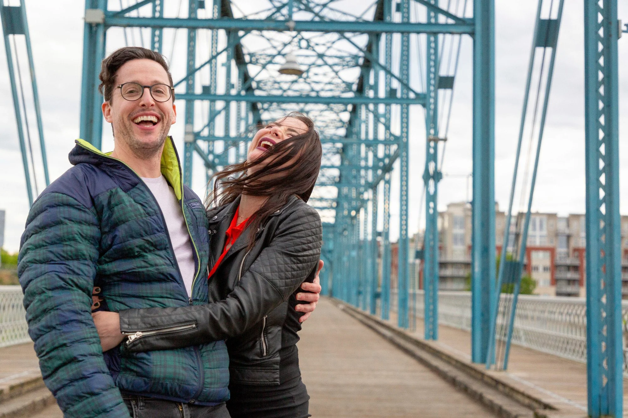 A smiling man and woman embrace on a bridge with blue metal framework, overcast sky in the background.