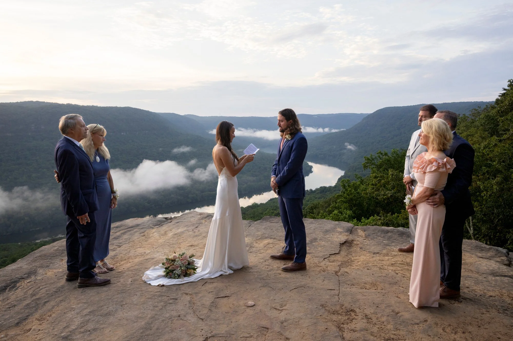 A wedding ceremony taking place outdoors on a rocky ledge with a river and forested mountains in the background. The bride and groom stand in the center, facing each other, with the bride reading vows. Six guests, three men and three women, dressed i