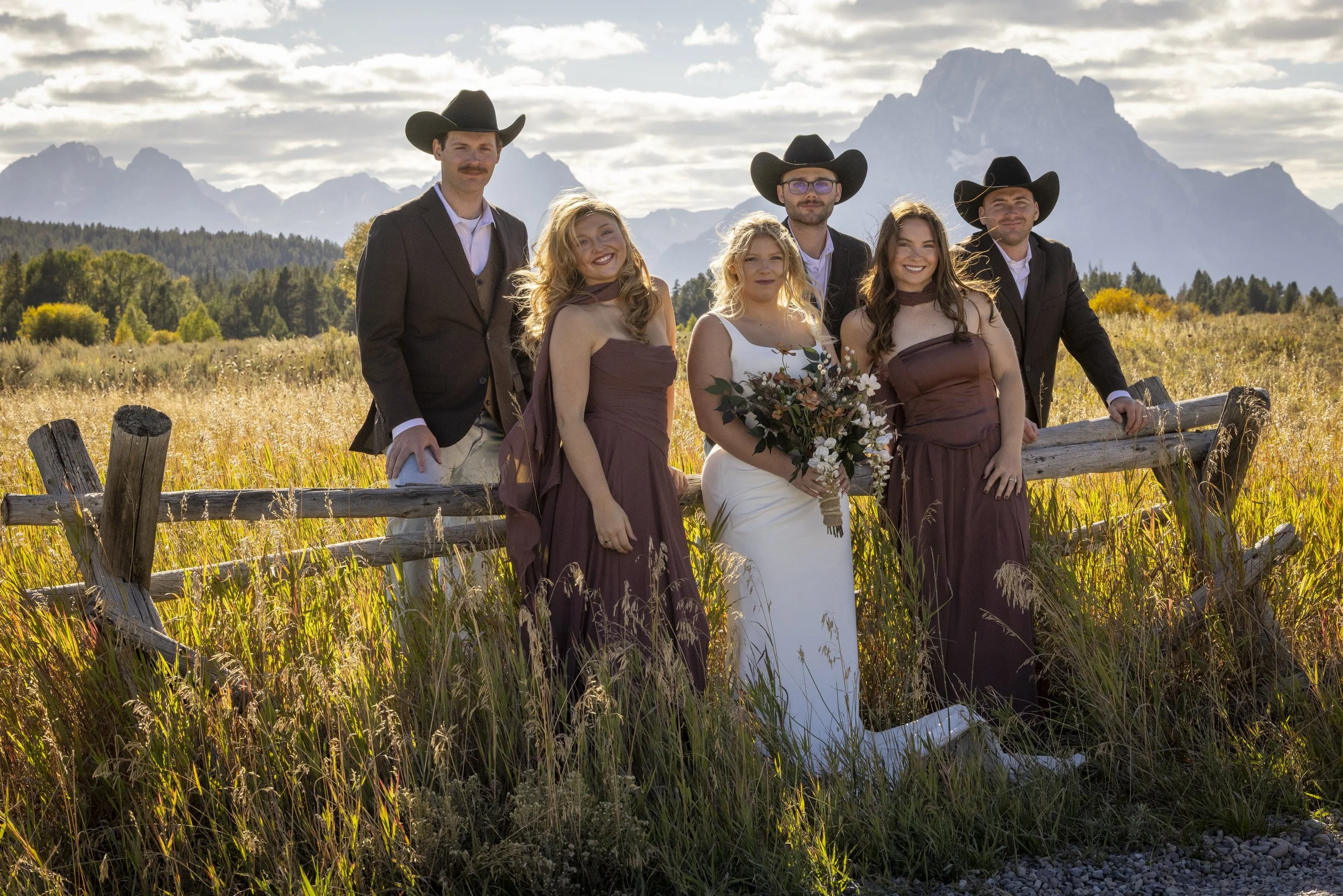 A group of six people, three men and three women, enjoying an outdoor wedding photo on a field with mountains in the background. The women are wearing dresses, and the men are in suits with cowboy hats.