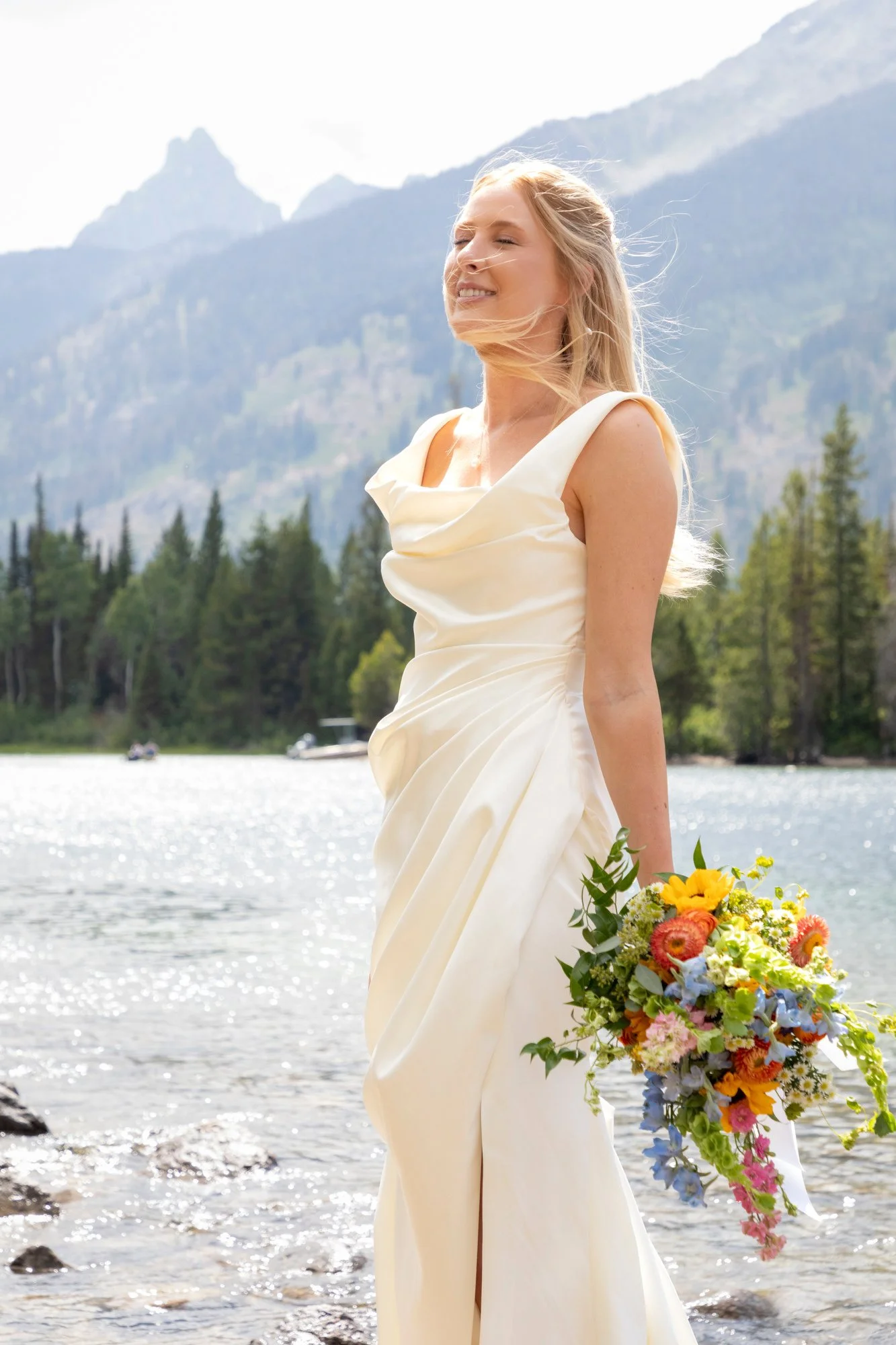A woman in a white wedding dress standing by a lake, holding a colorful bouquet of flowers, with mountains and trees in the background, her eyes closed and about to smile.