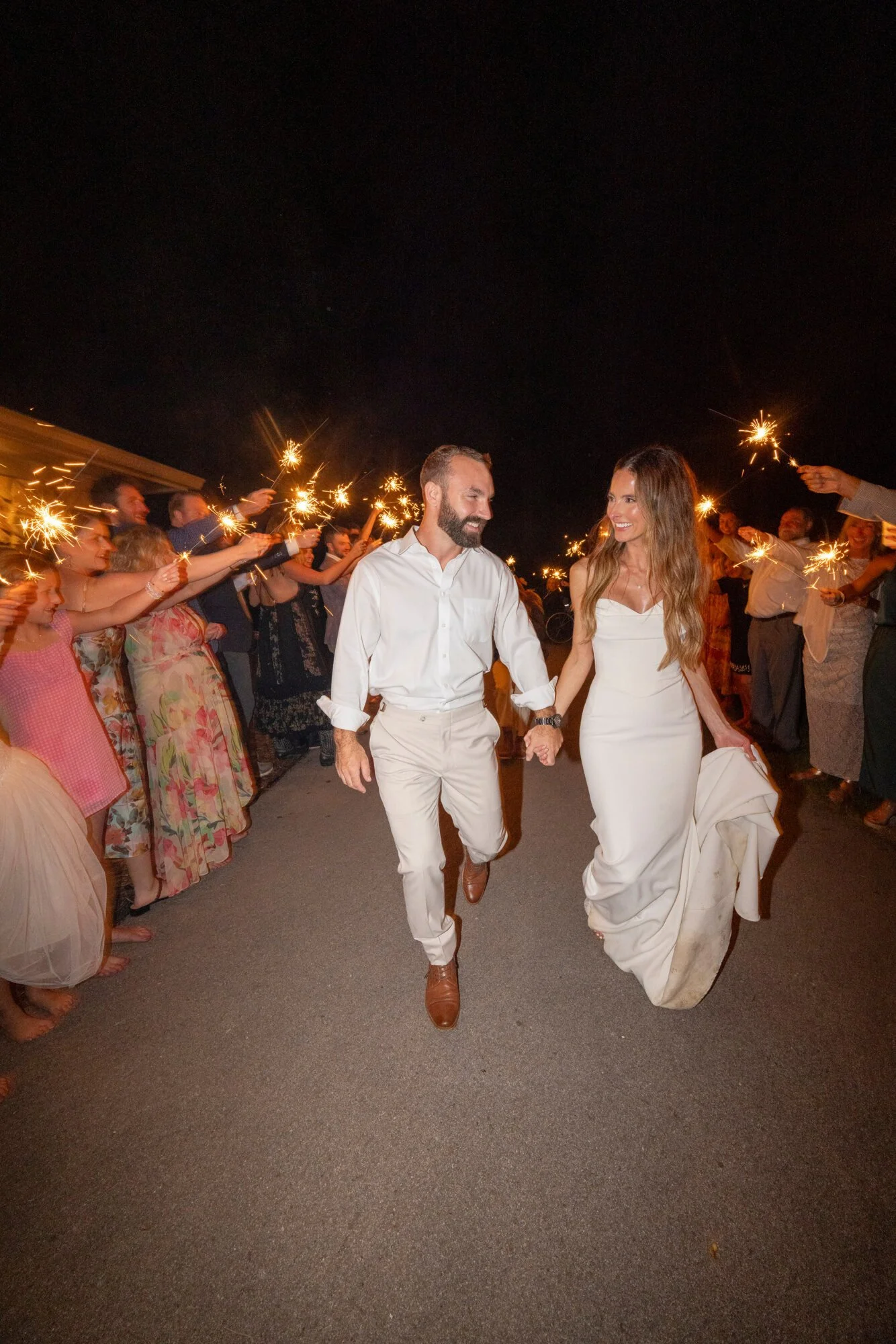 A bride and groom hold hands and run together at night while friends and family cheer and wave sparklers during a celebration.
