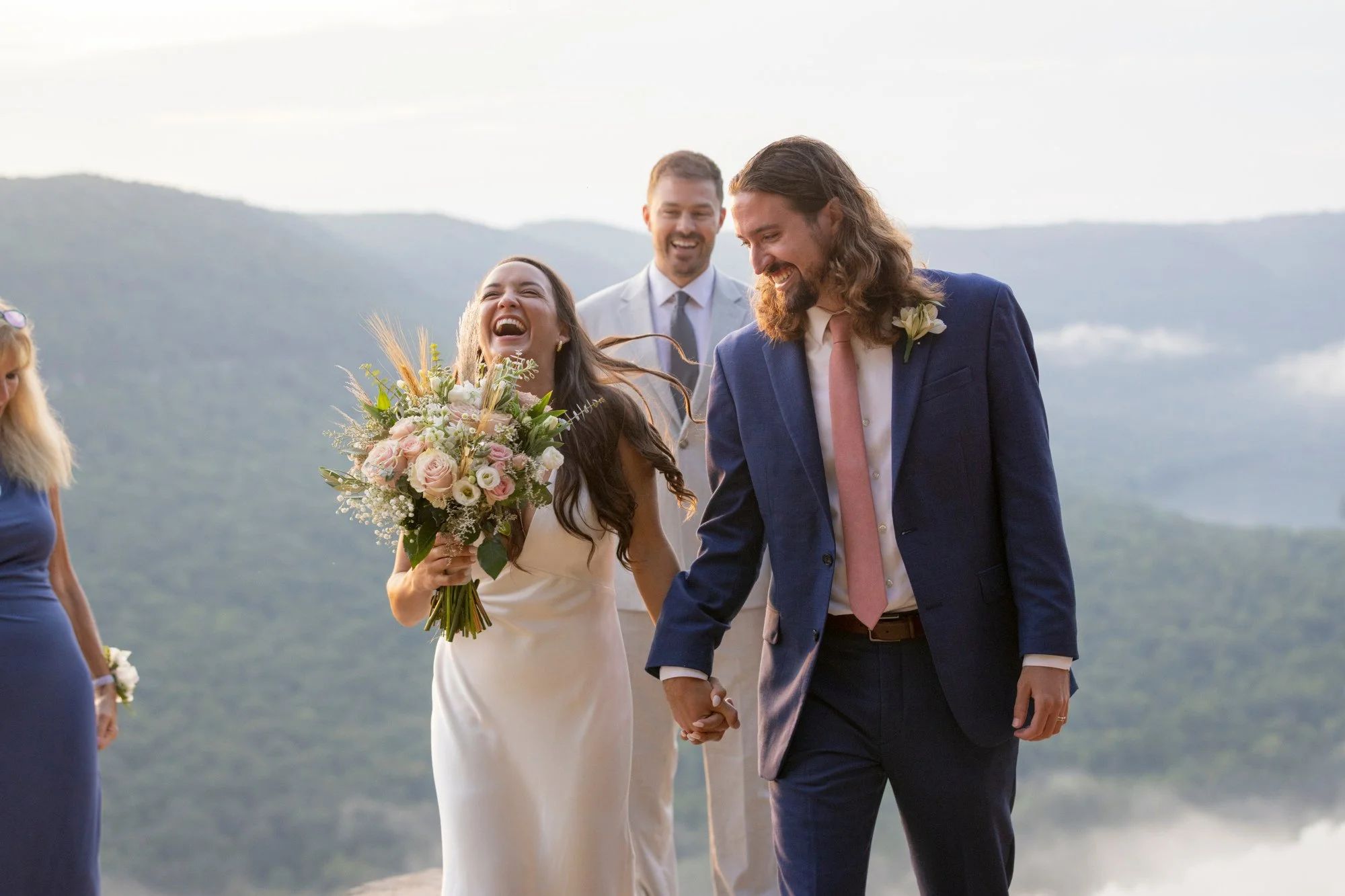 A couple is holding hands and walking outdoors during sunset, with the woman holding a bouquet of flowers and smiling, while the man also smiling, dressed in a blue suit with a pink tie, and there are other people nearby.