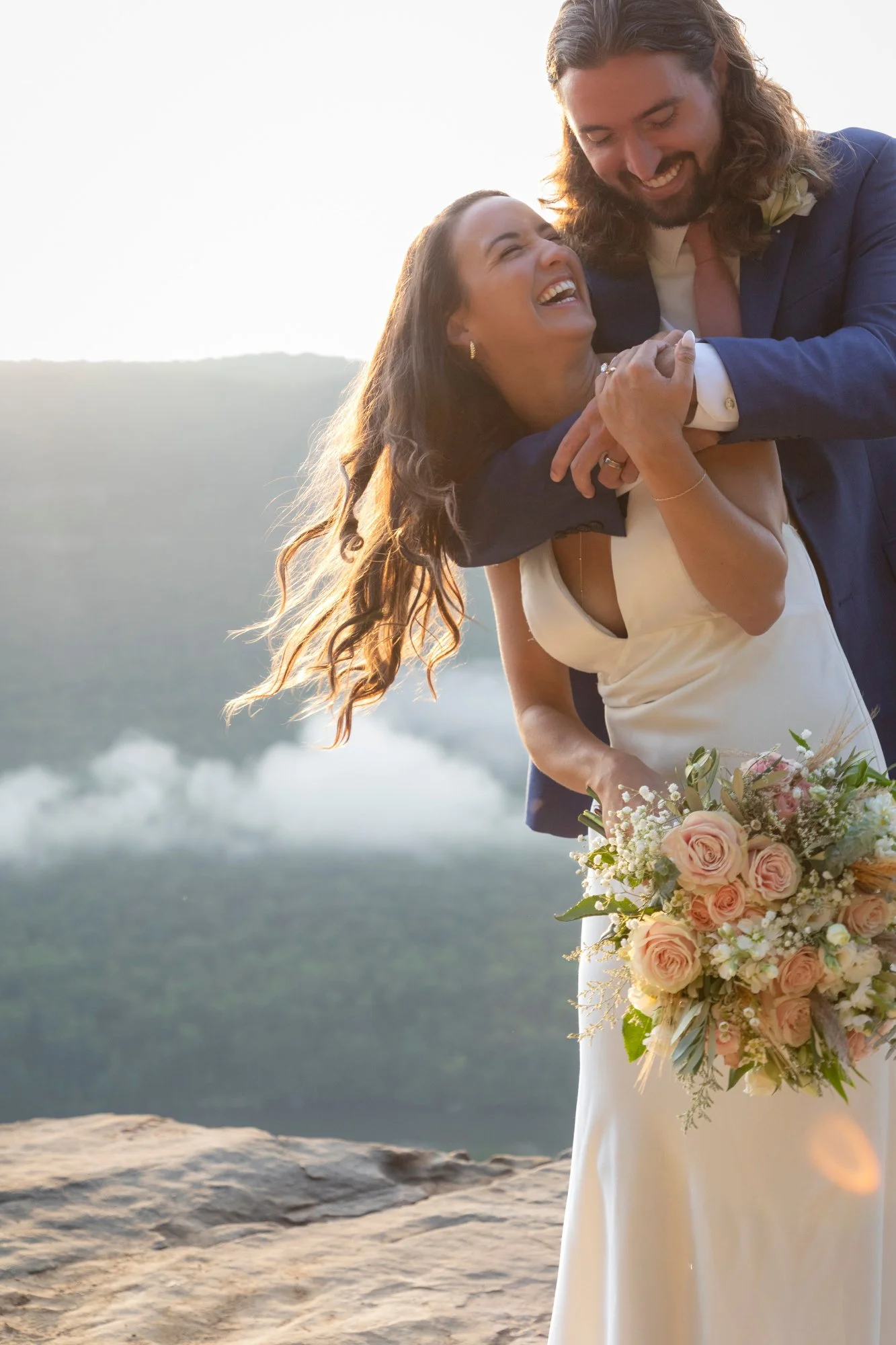 A smiling bride and groom hugging during their wedding on a rocky outdoor location with a scenic background of hills and a body of water, the bride holding a large bouquet of pink roses and white flowers.