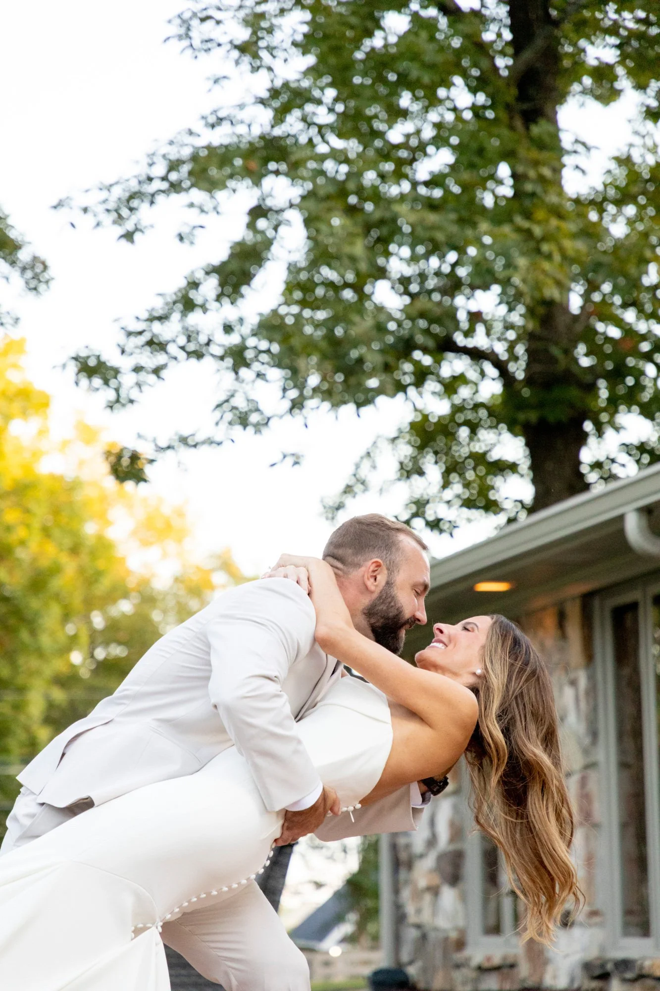 A couple dancing outdoors with a large green tree and a house in the background, enjoying a joyful moment together.