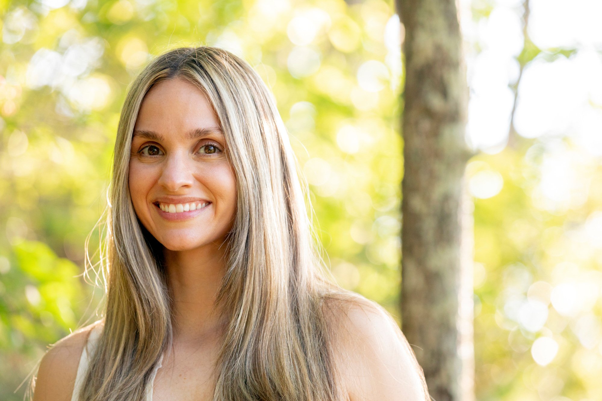 A young woman with long blonde hair outdoors, smiling, with a background of green trees and sunlight filtering through leaves.