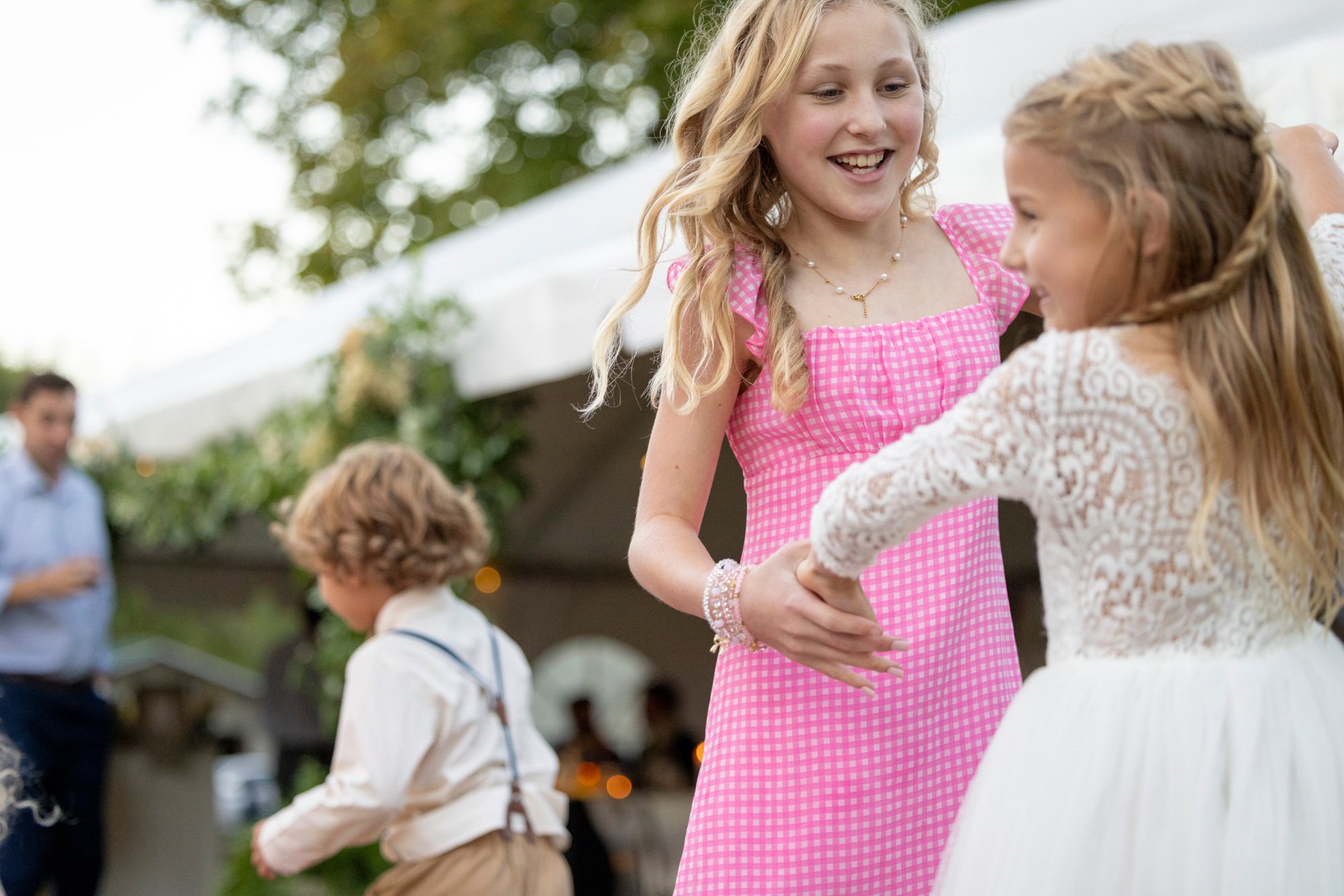 Two young girls with blonde hair smiling and dancing at an outdoor party, one wearing a pink checkered dress and the other wearing a white lace dress. Other children and adults are visible in the background, with some holding glasses.