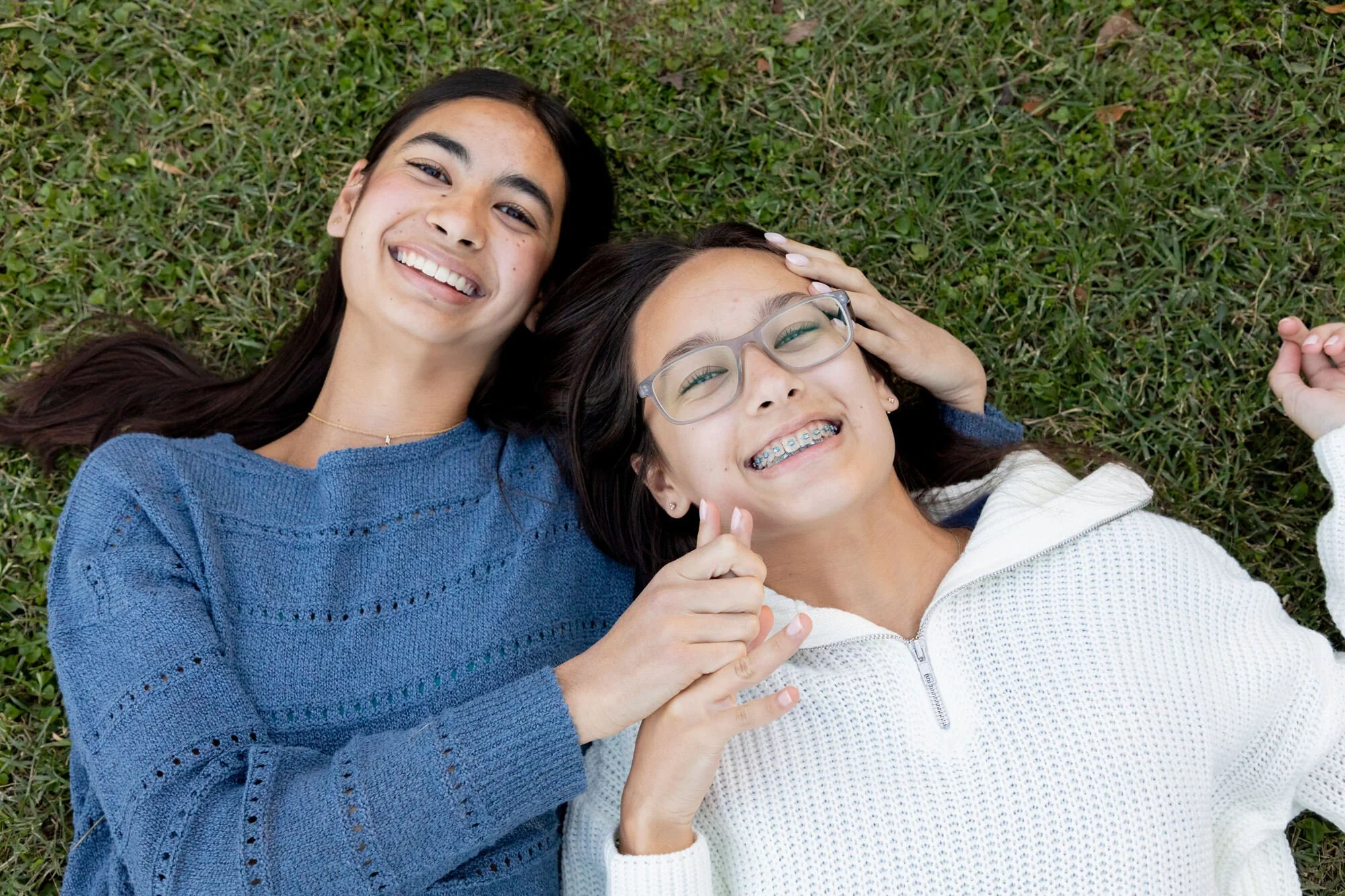 Two young women lying on grass and smiling, with one wearing glasses and braces and the other with long brown hair, both wearing sweaters.