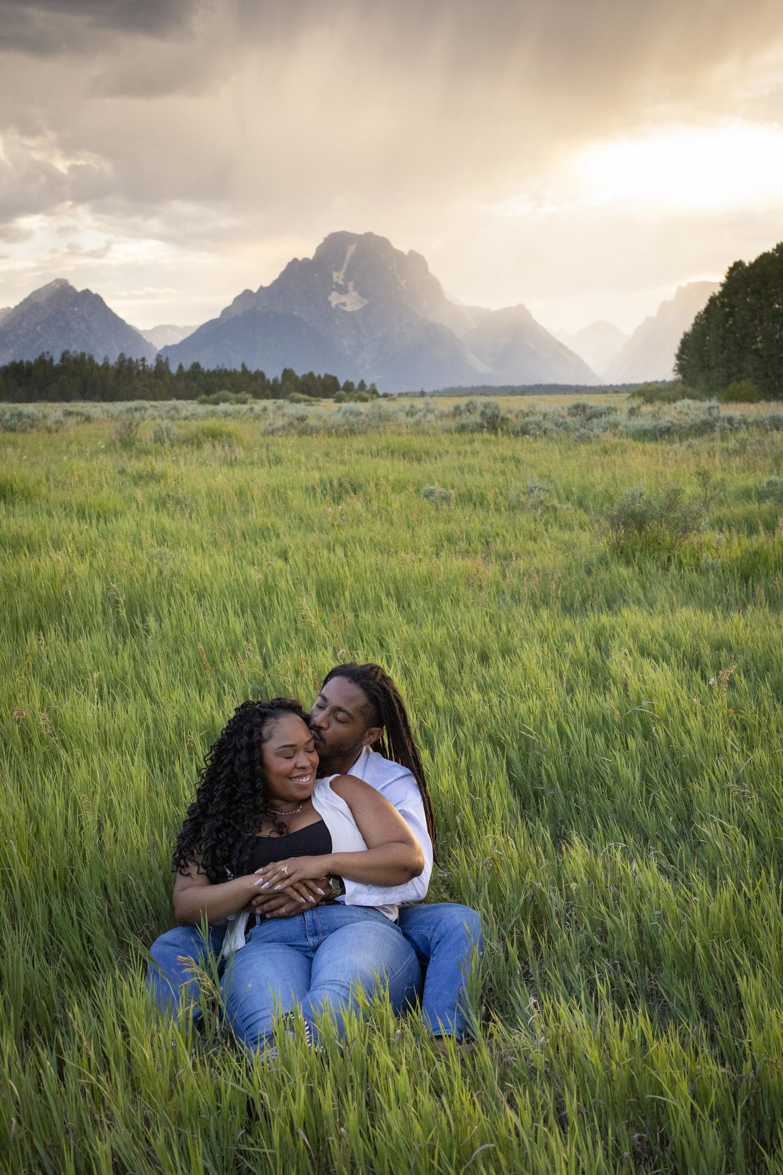 A couple hugging and smiling in a grassy field with mountains in the background during sunset.