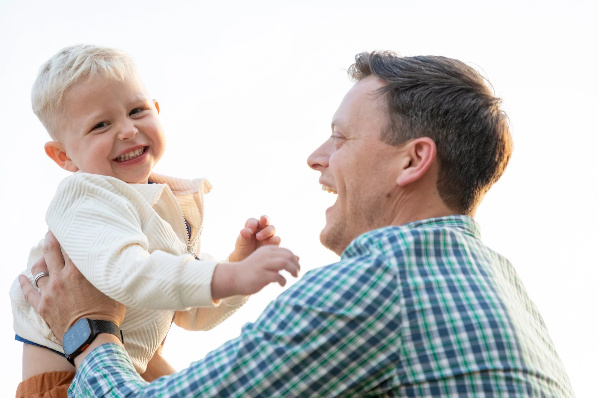 A man with dark hair, wearing a plaid shirt, holding a young boy with blonde hair, cotton jacket, and orange pants. They are smiling and looking at each other outdoors against a clear sky.