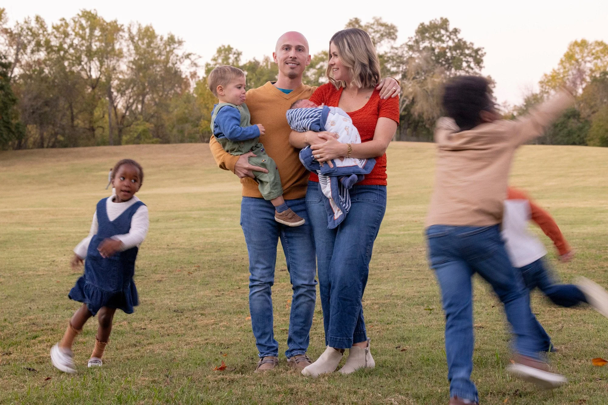 A family with two children standing on a grassy field, with Trees in the background. The mother is holding a baby, while the father has a young boy on his arm. A girl is running towards the family.