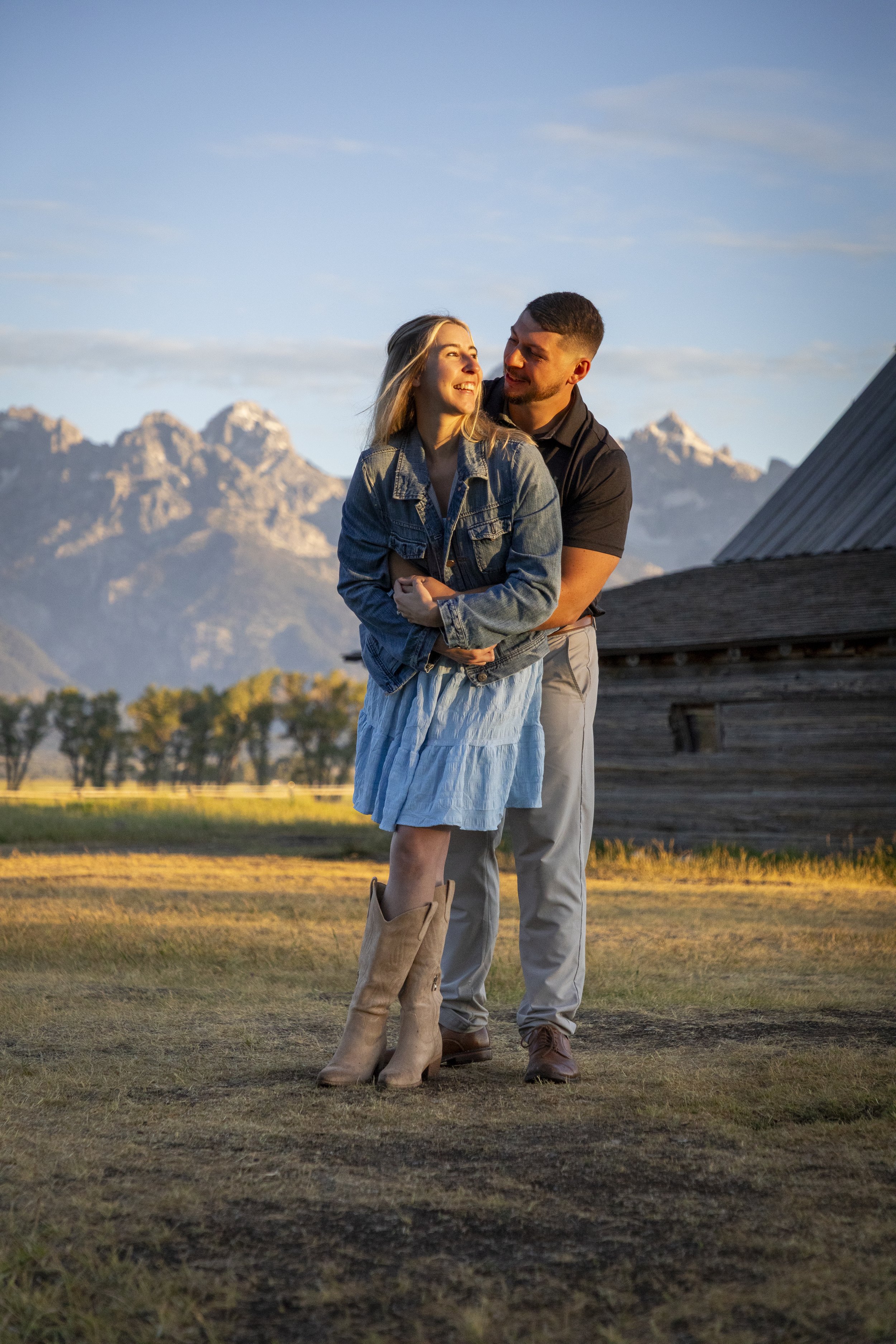 A couple standing outdoors during sunset with mountains in the background, the woman wearing a denim jacket and a light blue skirt, and the man hugging her from behind, both smiling.