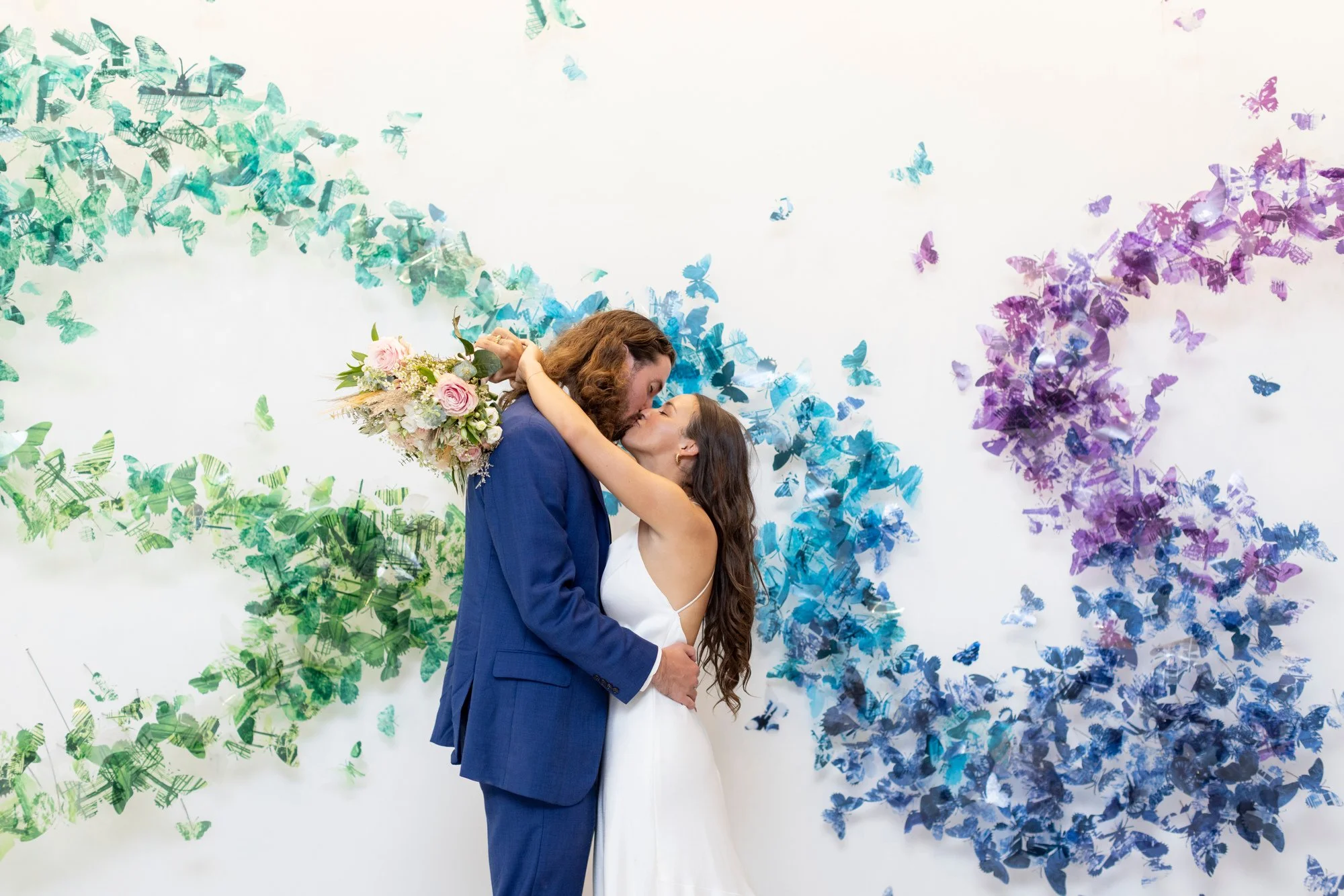 A couple hugging and kissing at their wedding, holding a bouquet of flowers with colorful butterflies on a white background.