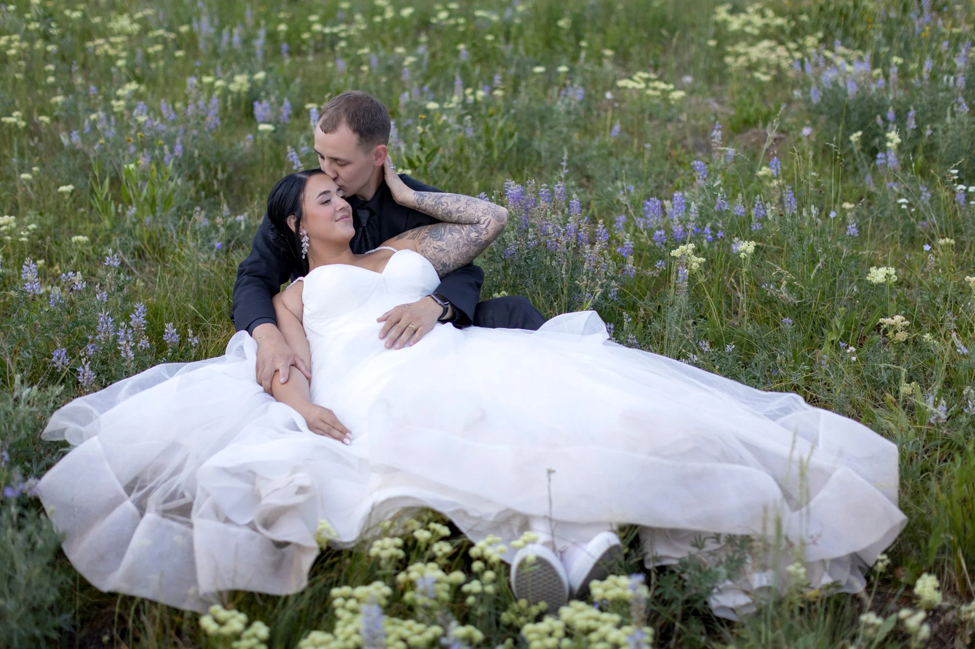 A man and woman lying in a field of wildflowers, with the man kissing the woman's forehead. The woman is wearing a white wedding dress and earrings, and the man is dressed in black with tattoos visible on his arm.