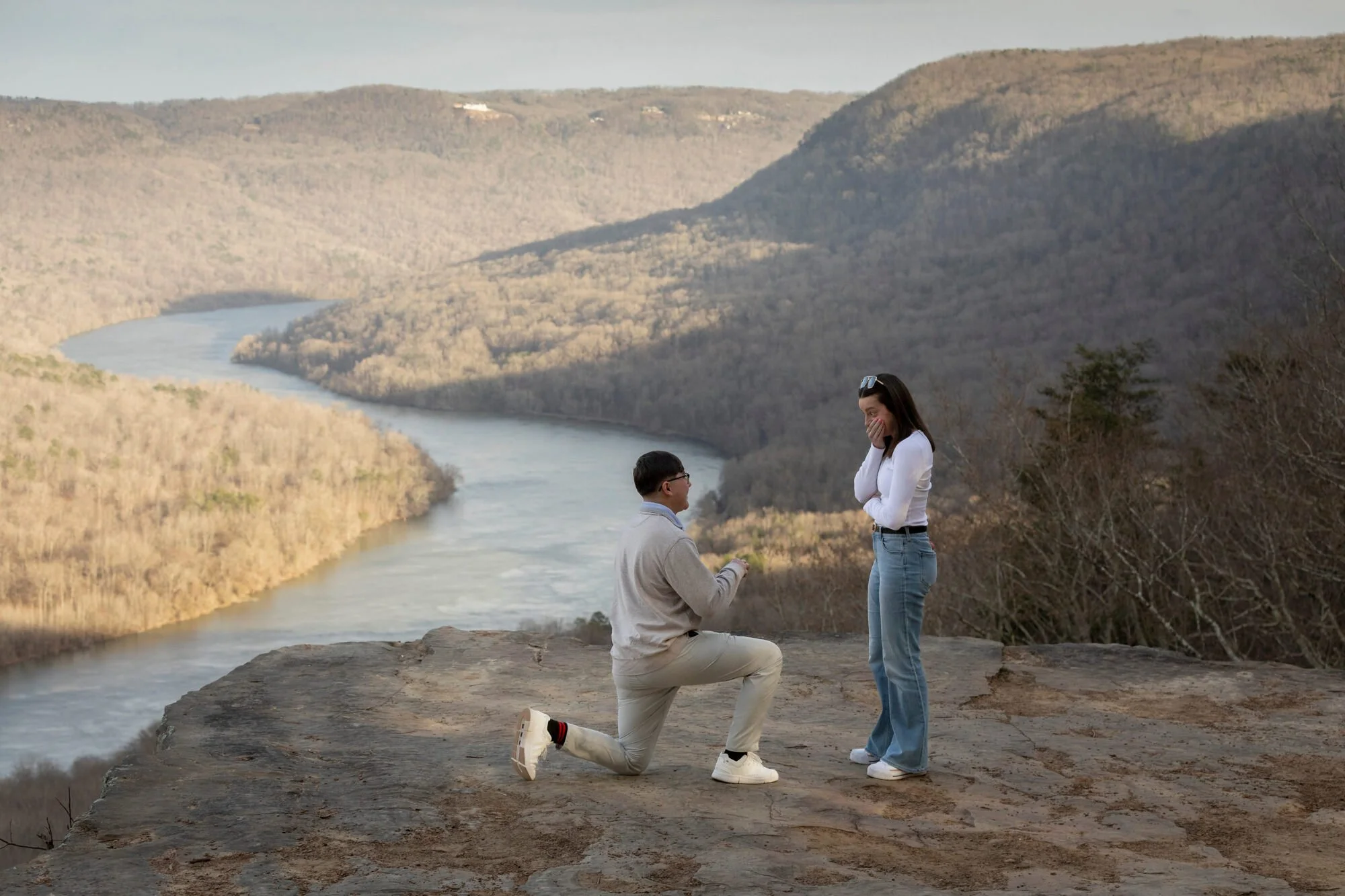 A couple is engaged in a marriage proposal on a rocky overlook near a river, with forested hills in the background. The man is kneeling on one knee holding out a ring, while the woman stands with her hand covering her mouth, both smiling. The scenery