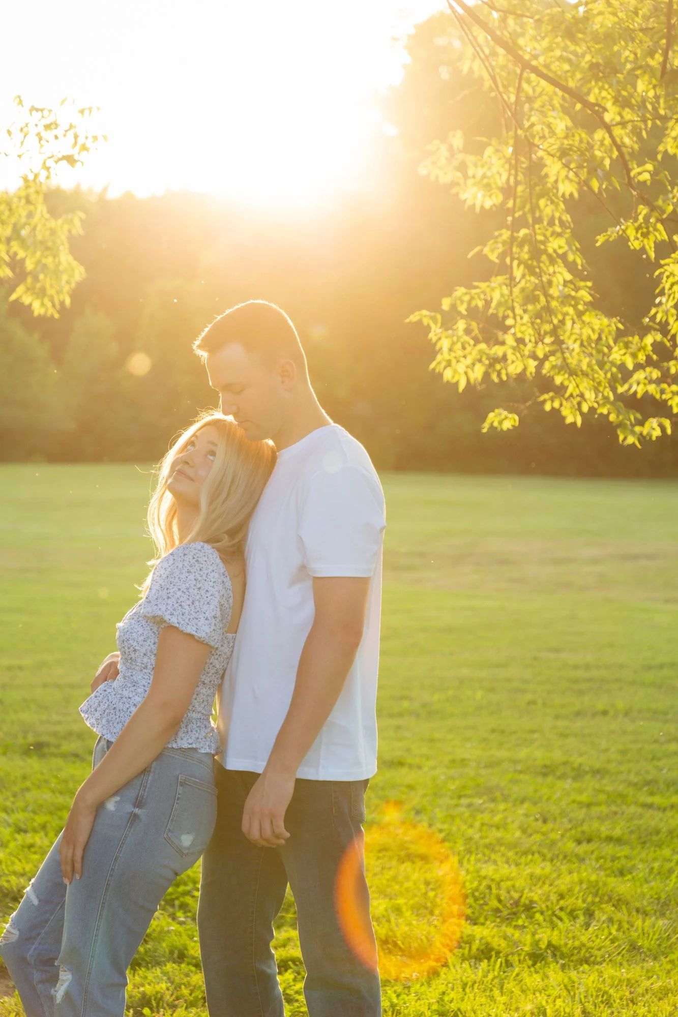 A young couple standing outdoors in a grassy field with trees, backlit by the setting sun, creating a warm, golden glow.
