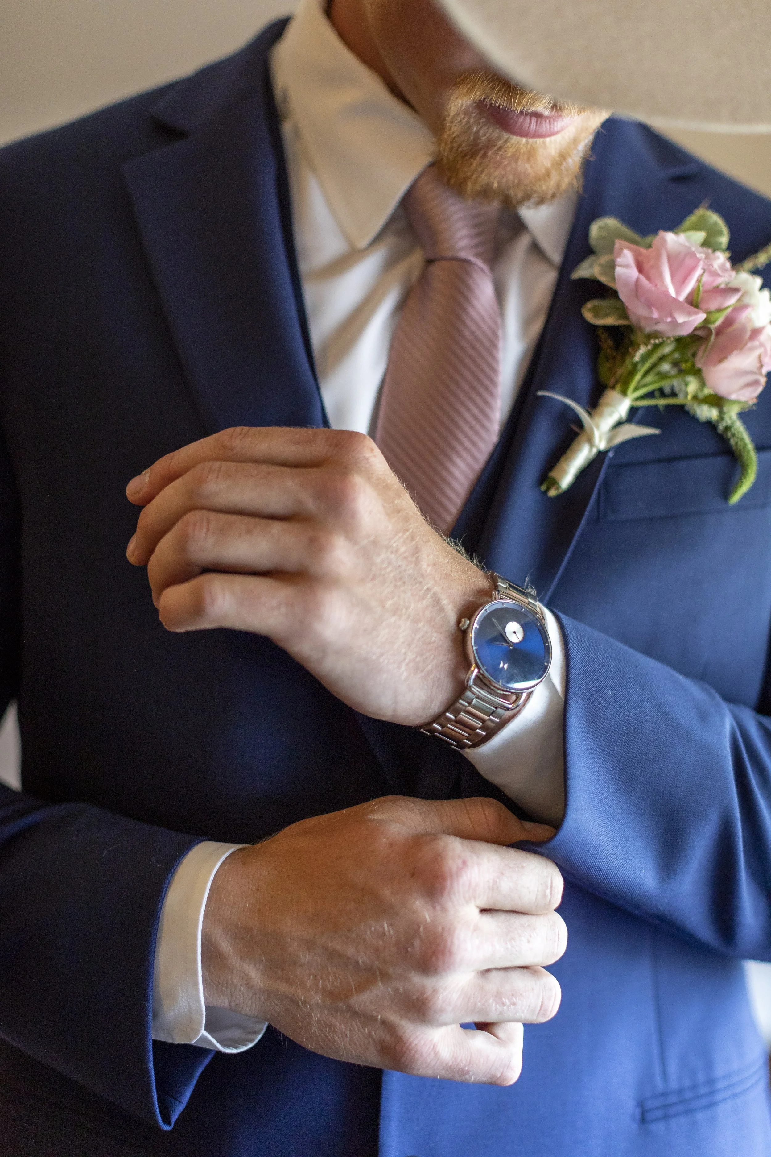 Close-up of a man in a blue suit with a pink tie and boutonniere, adjusting his watch on his wrist.