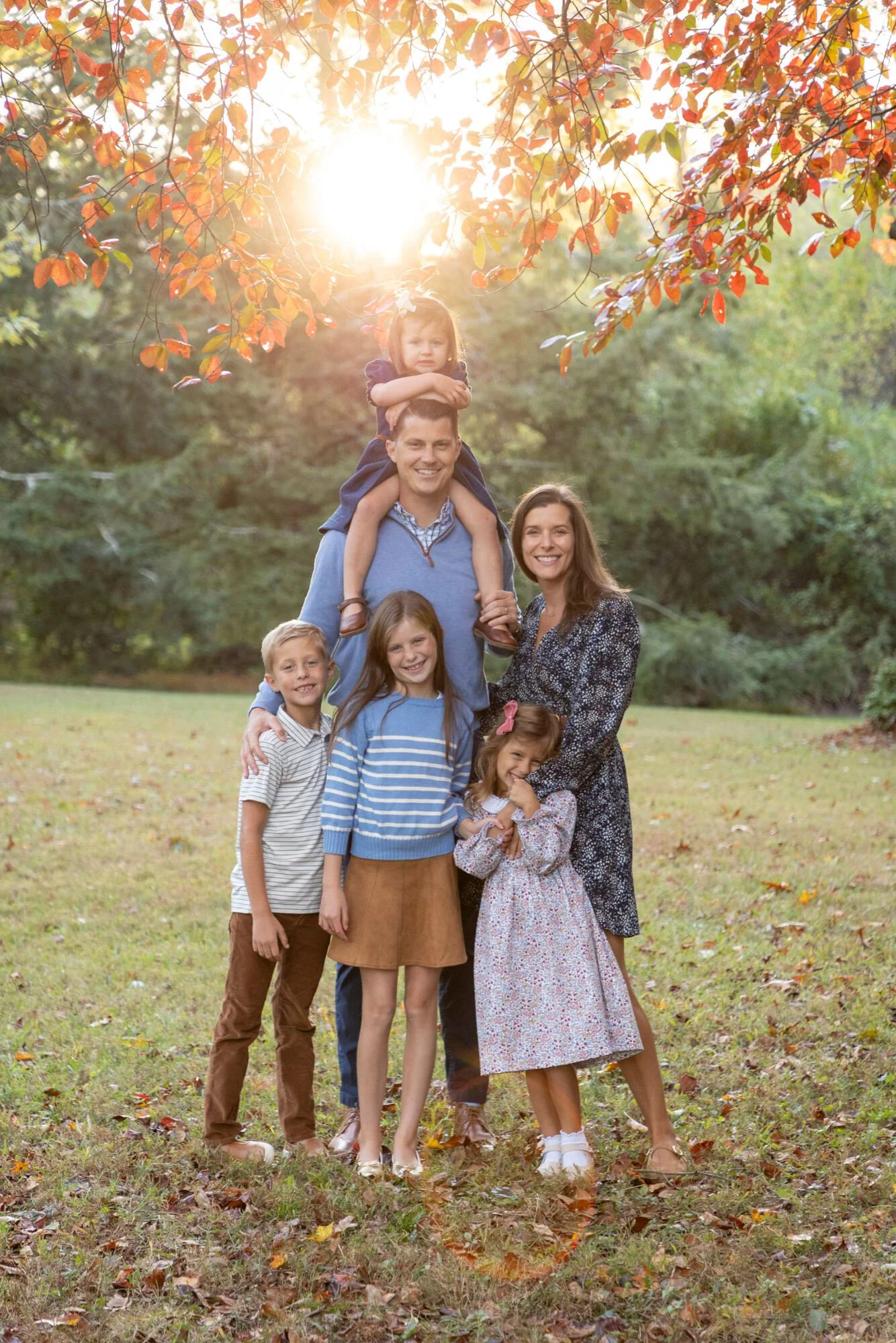 A family of six outdoors during autumn, with children and adults smiling, one child riding on a man's shoulders, all standing on grass with fallen leaves, trees with orange and green leaves, backlit by sunlight.