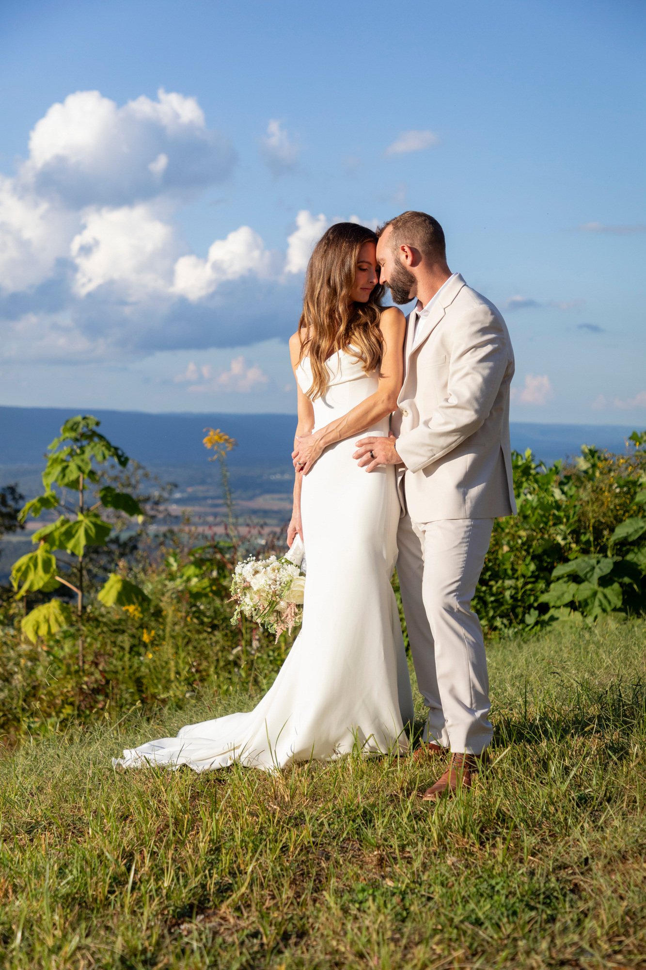 A bride and groom stand closely together outdoors on a grassy hill, with a scenic landscape of mountains, trees, and a bright blue sky with fluffy clouds in the background. The bride wears a white wedding gown and holds a bouquet, while the groom is 