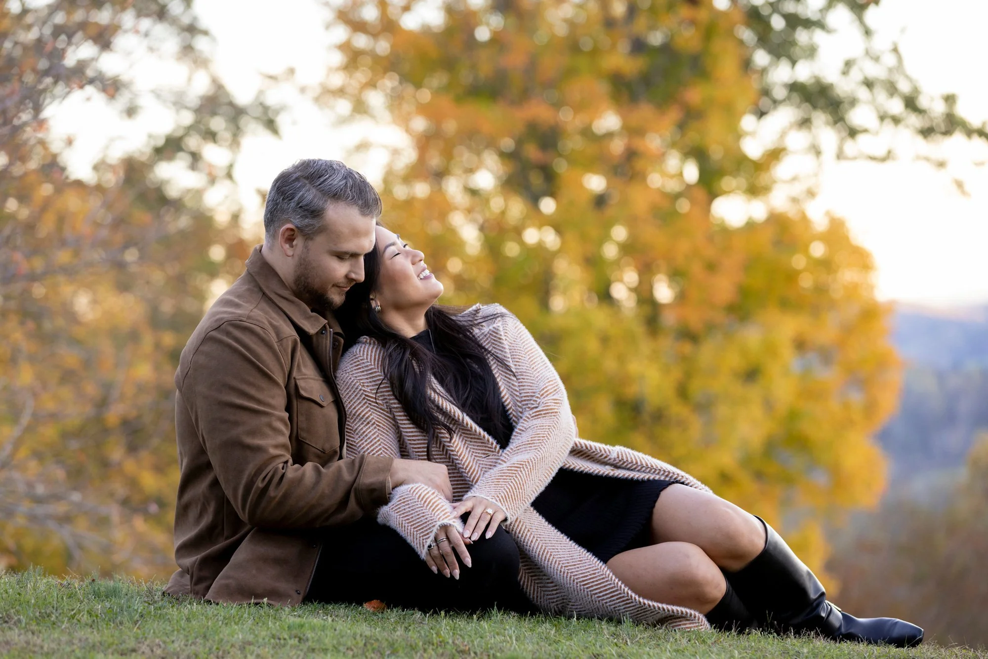 A couple sitting on the grass during autumn, with the woman smiling and leaning back as the man holds her. They are outdoors with colorful fall trees in the background.