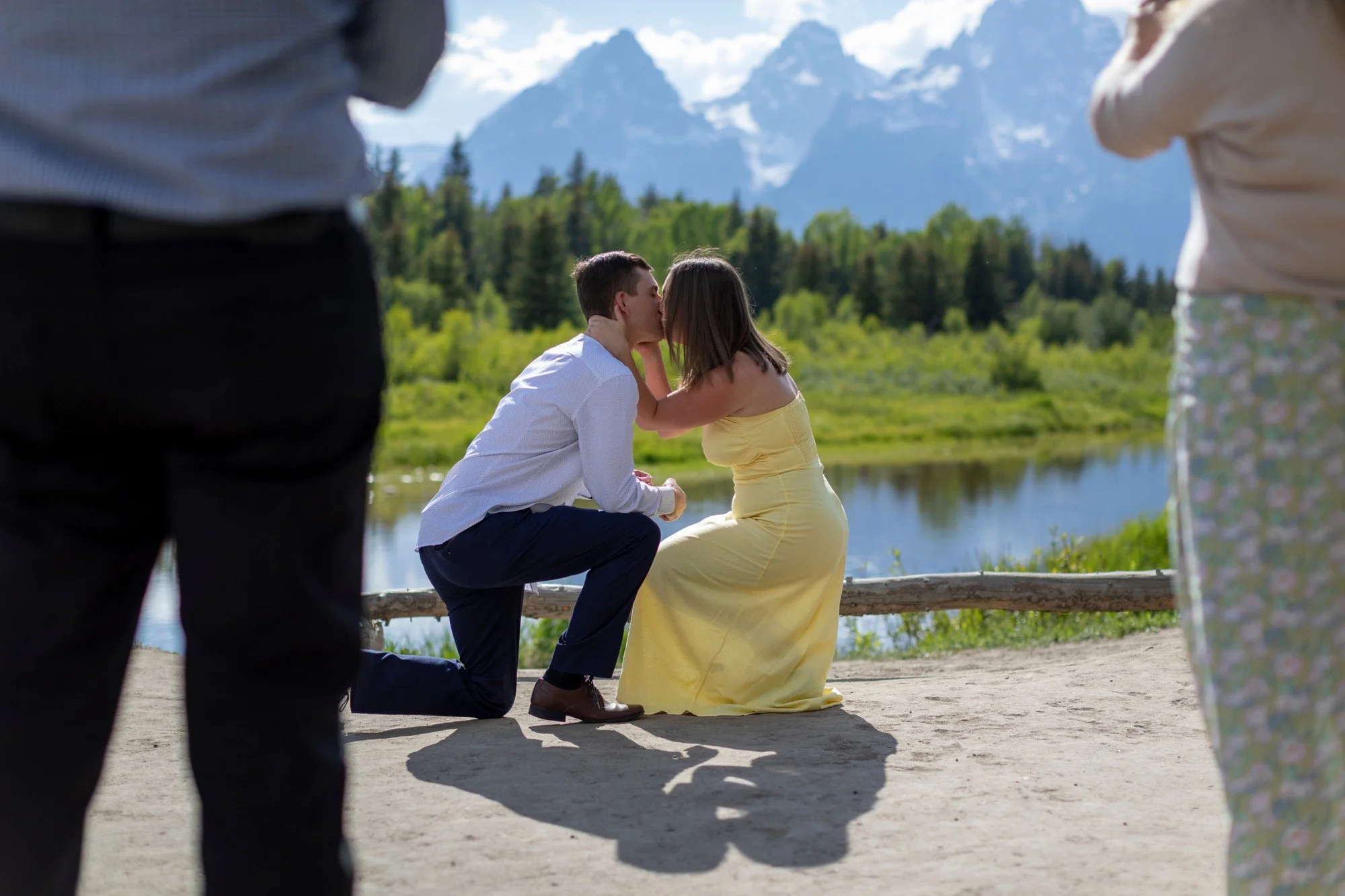 A couple is exchanging a kiss during an outdoor wedding ceremony by a river, with mountains and trees in the background. The bride is wearing a yellow dress, and the groom is kneeling in front of her. Two people are standing on either side, partially