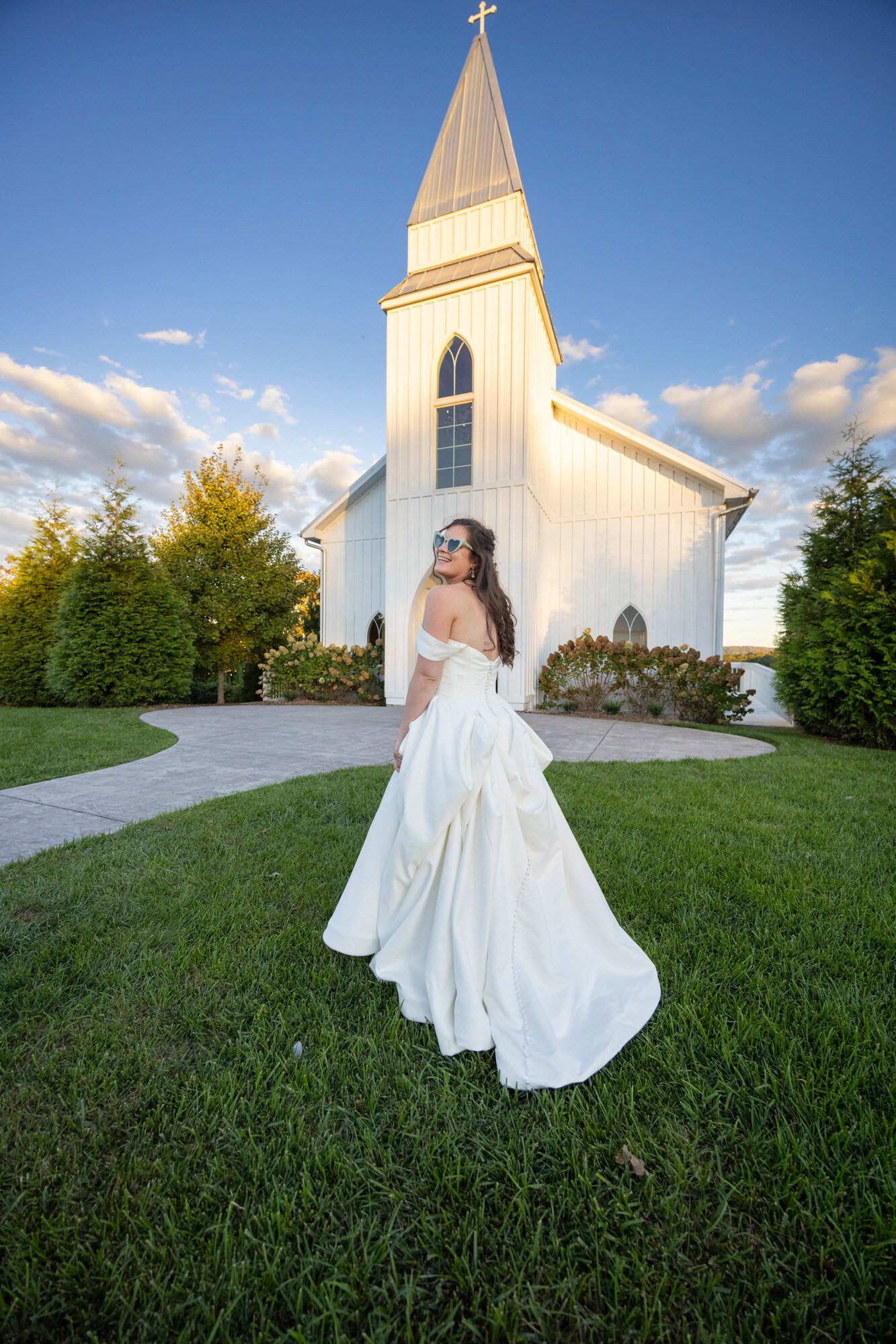 A woman in a wedding dress and heart-shaped sunglasses smiling while standing on a grassy lawn in front of a white church with a tall steeple during sunset.