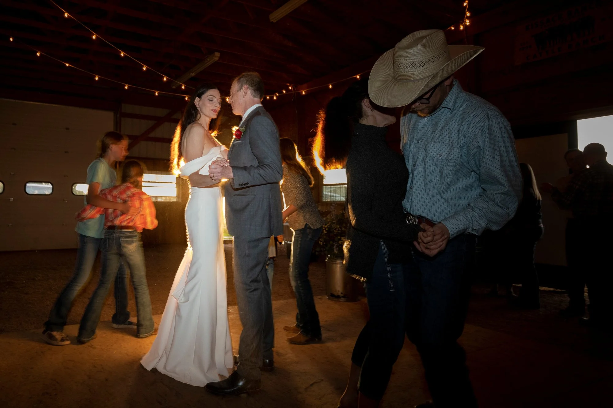 A couple dancing closely together in a barn with string lights, while other people dance and socialize in the background.