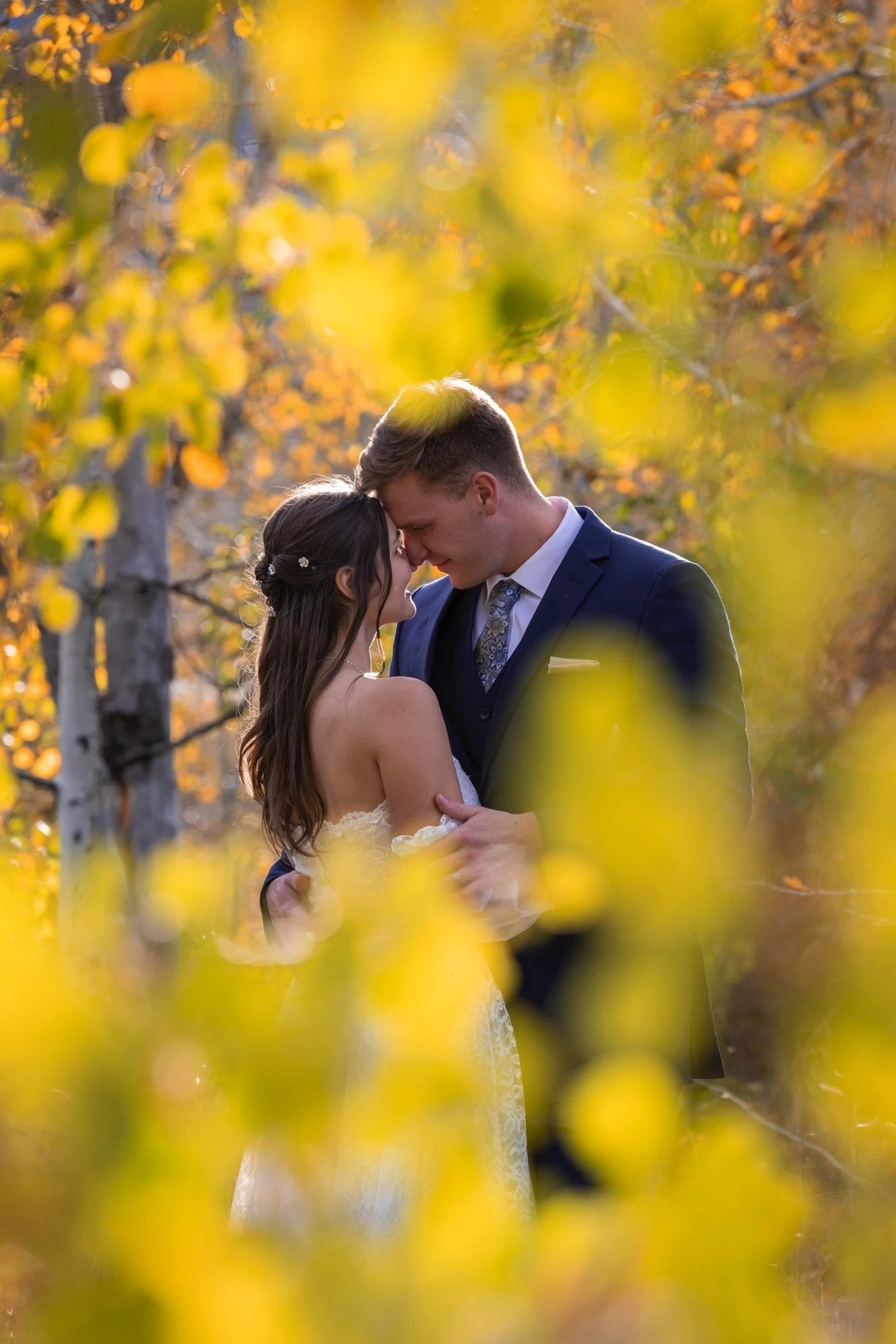A bride and groom embrace and touch foreheads in an outdoor setting with autumn leaves.