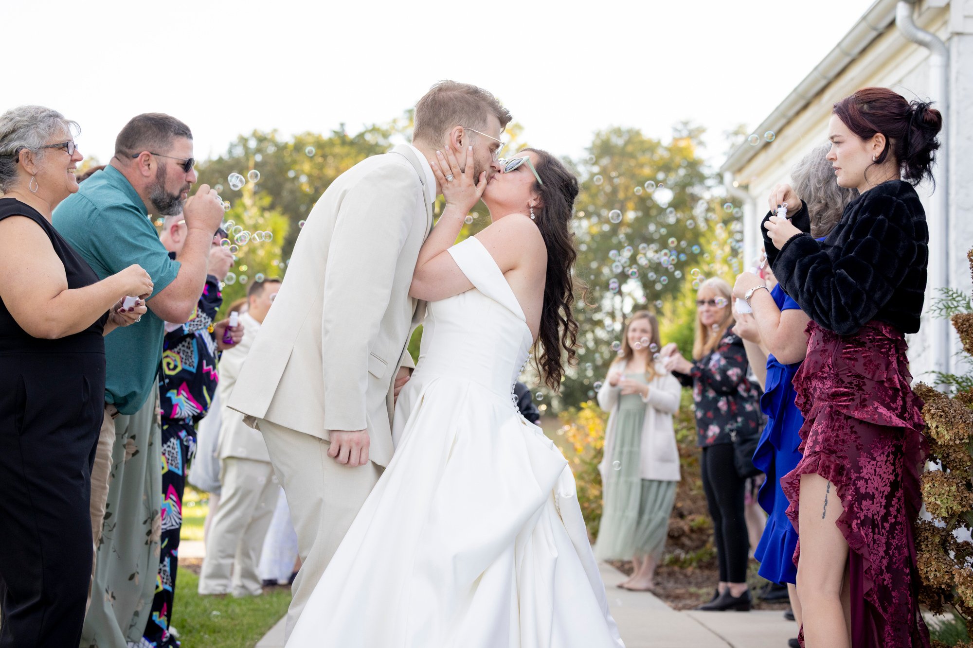 A bride and groom kiss during their outdoor wedding ceremony on a sunny day, surrounded by friends and family, some blowing bubbles.