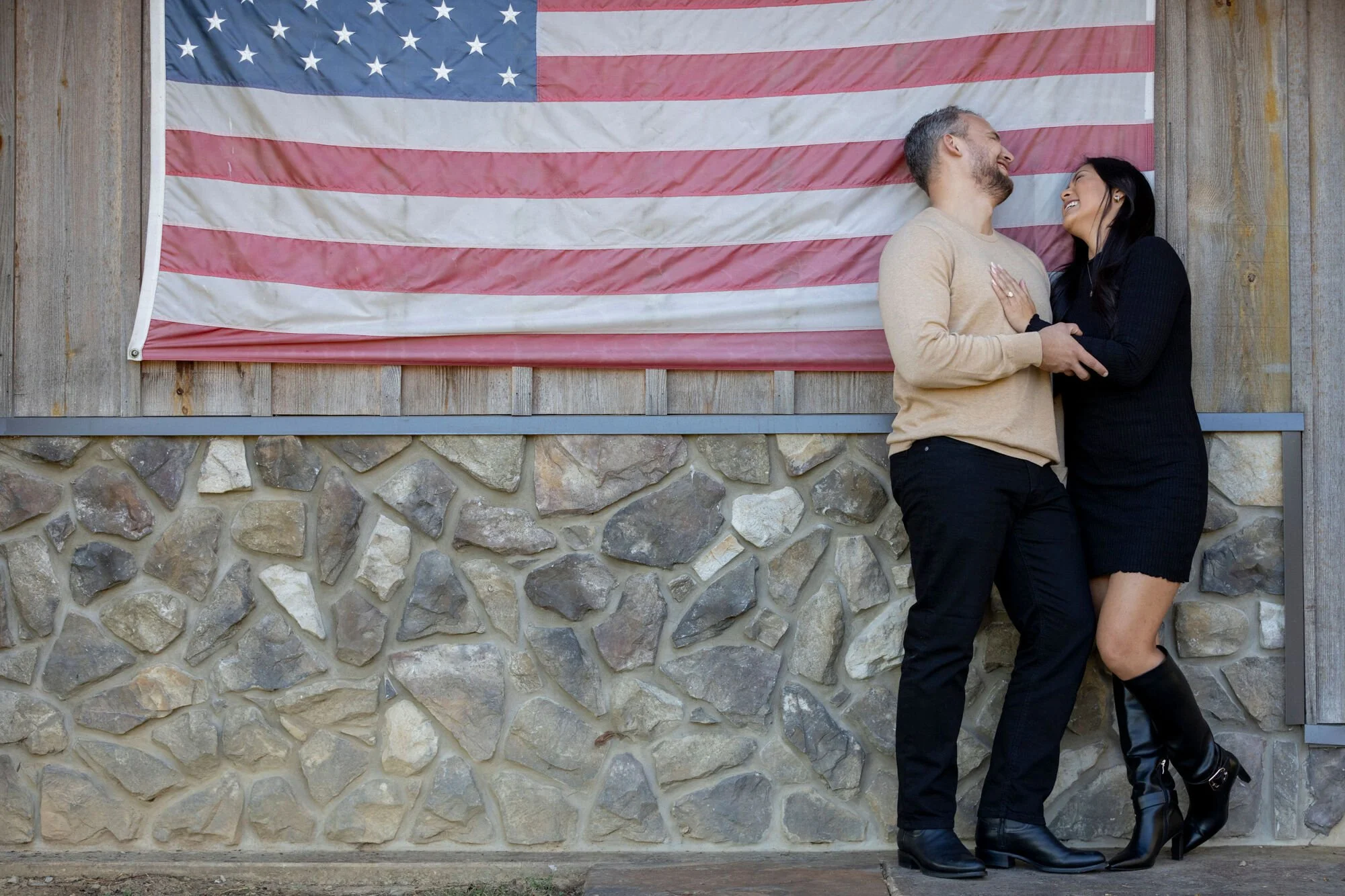 A couple standing close together in front of a large American flag, smiling and leaning her head on his shoulder.