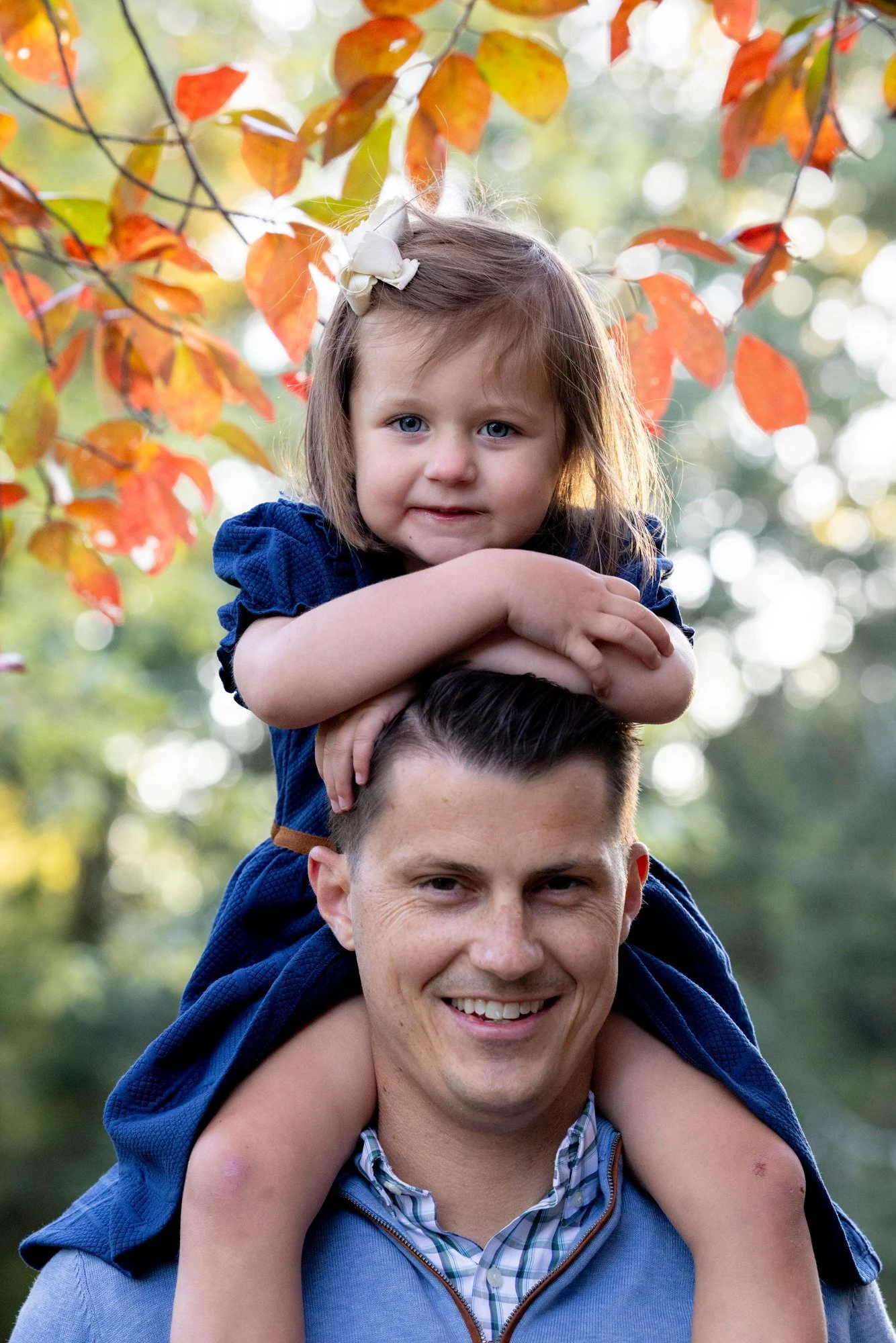 A man carrying a young girl on his shoulders in a park with autumn-colored leaves in the background.