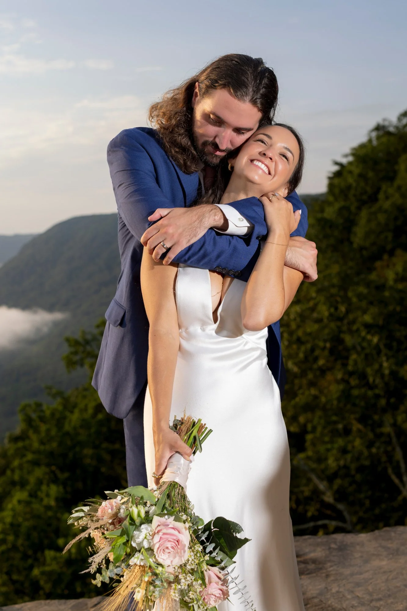 A bride and groom embrace outdoors in a scenic mountain setting. The bride is holding a large bouquet of pink and white flowers, wearing a satin wedding gown, and smiling. The groom is dressed in a blue suit, has long curly hair and a beard, and is l