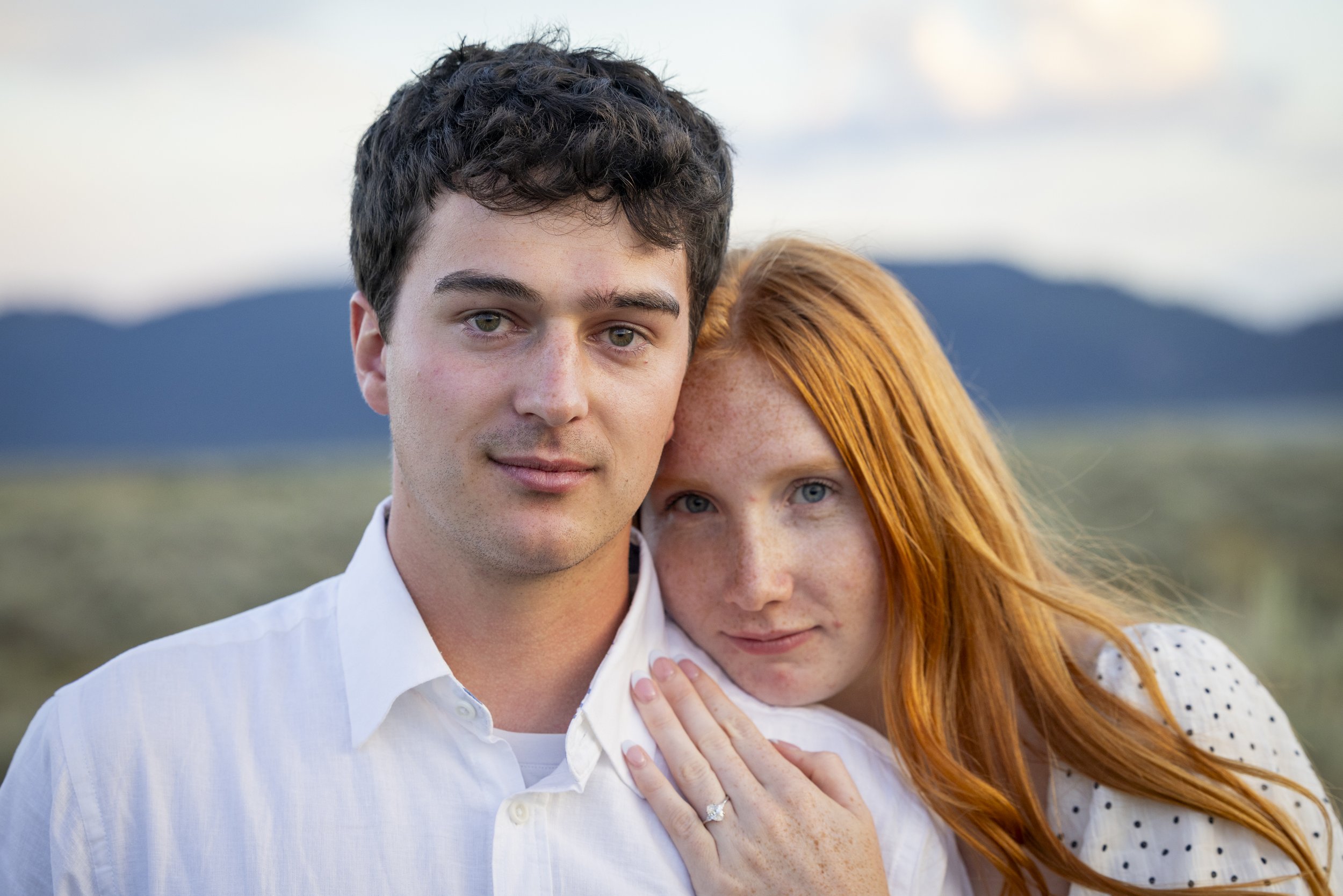 Close-up of a young man and woman standing closely outdoors, with mountains and sky in the background.