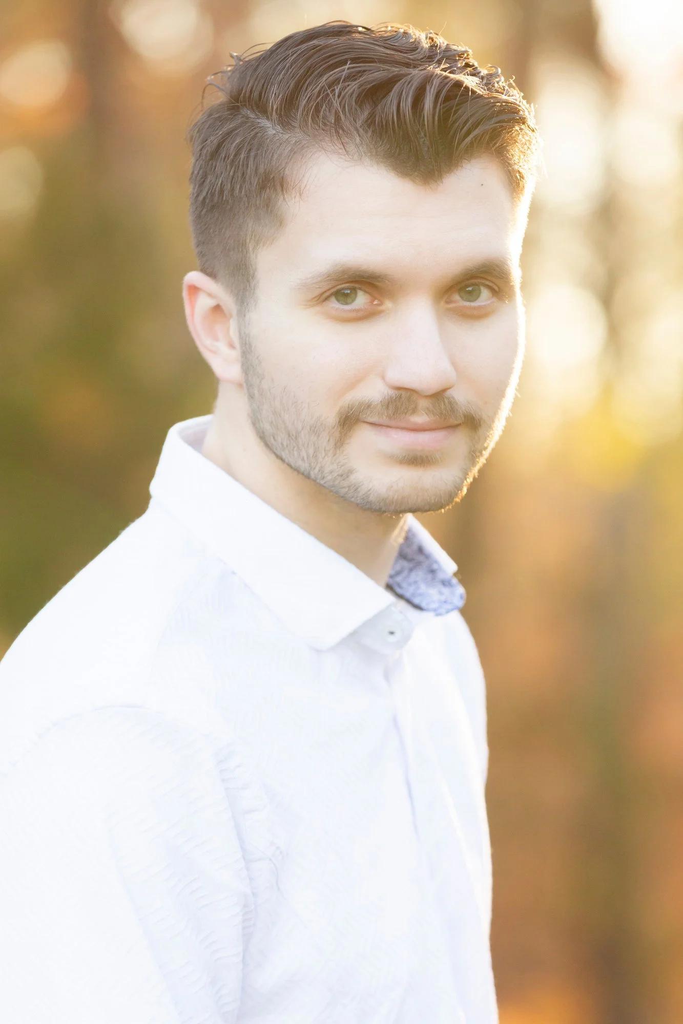 A young man with short, styled brown hair and green eyes, wearing a white collared shirt, stands outdoors in soft, golden sunlight with a blurred background of trees.