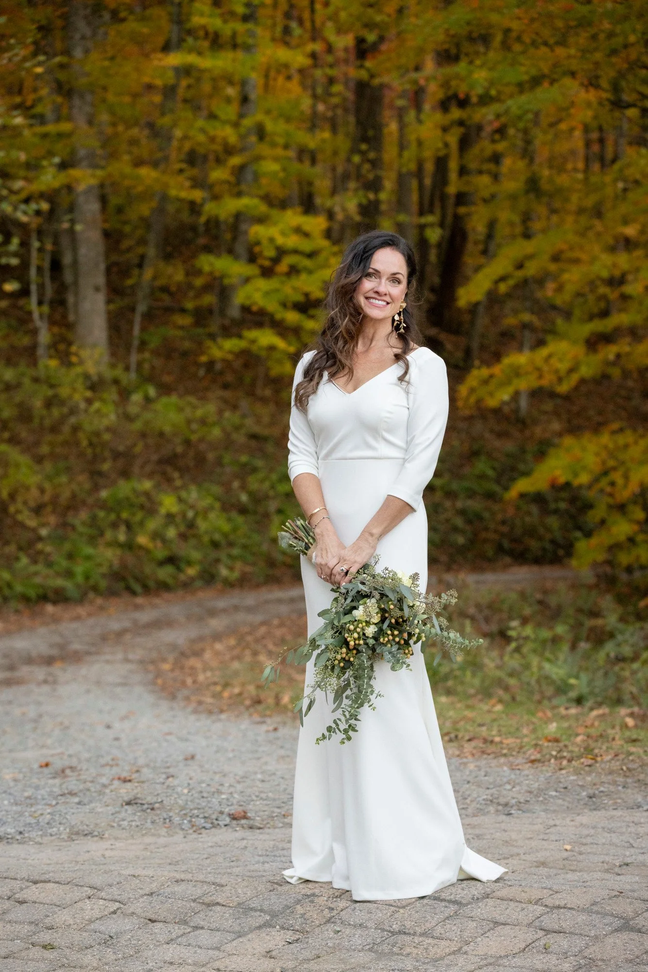 A woman in a white dress holding a bouquet of greenery, standing on a gravel path in front of a forest with autumn-colored leaves.