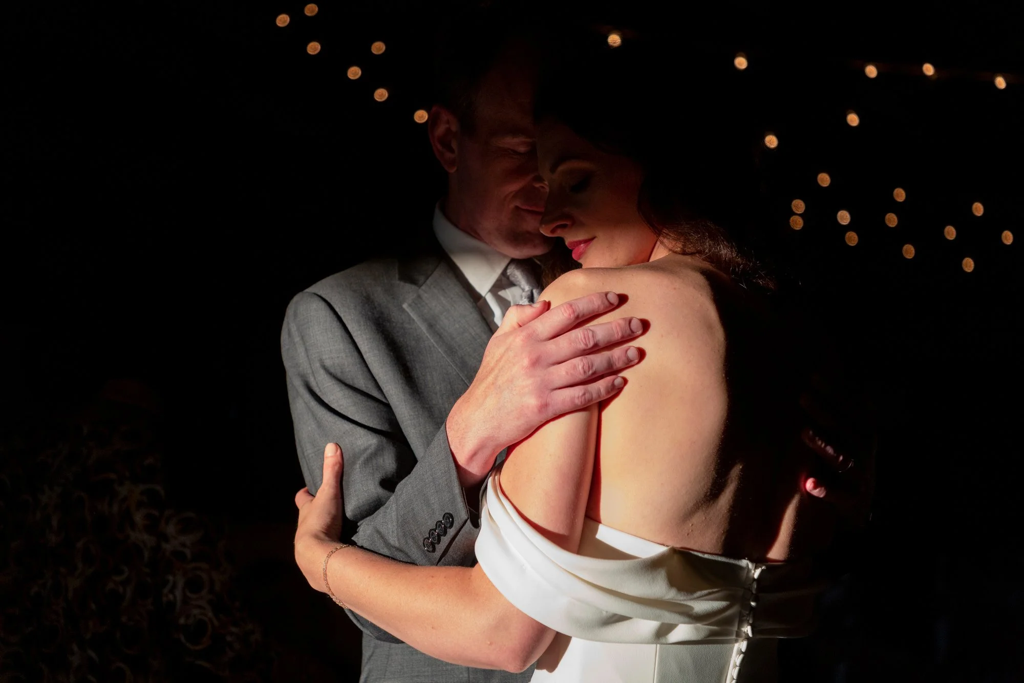 A man and woman embrace each other closely, with their heads touching and eyes closed, in a dark setting with string lights in the background.