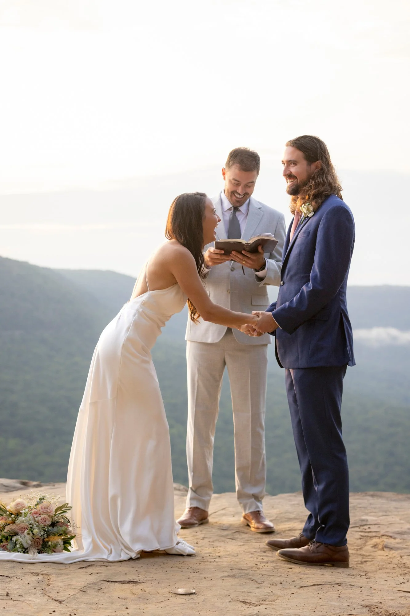 A wedding ceremony taking place outdoors with a bride and groom holding hands, an officiant reading from a book, and a scenic mountain landscape in the background.