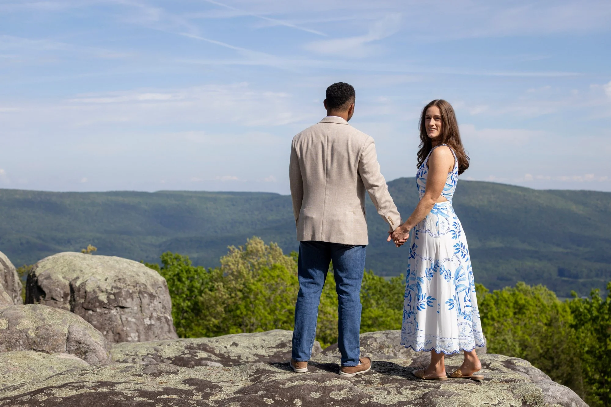 A couple holding hands and standing on rocks, facing each other in a scenic outdoor setting with green mountains and a blue sky with wispy clouds in the background.