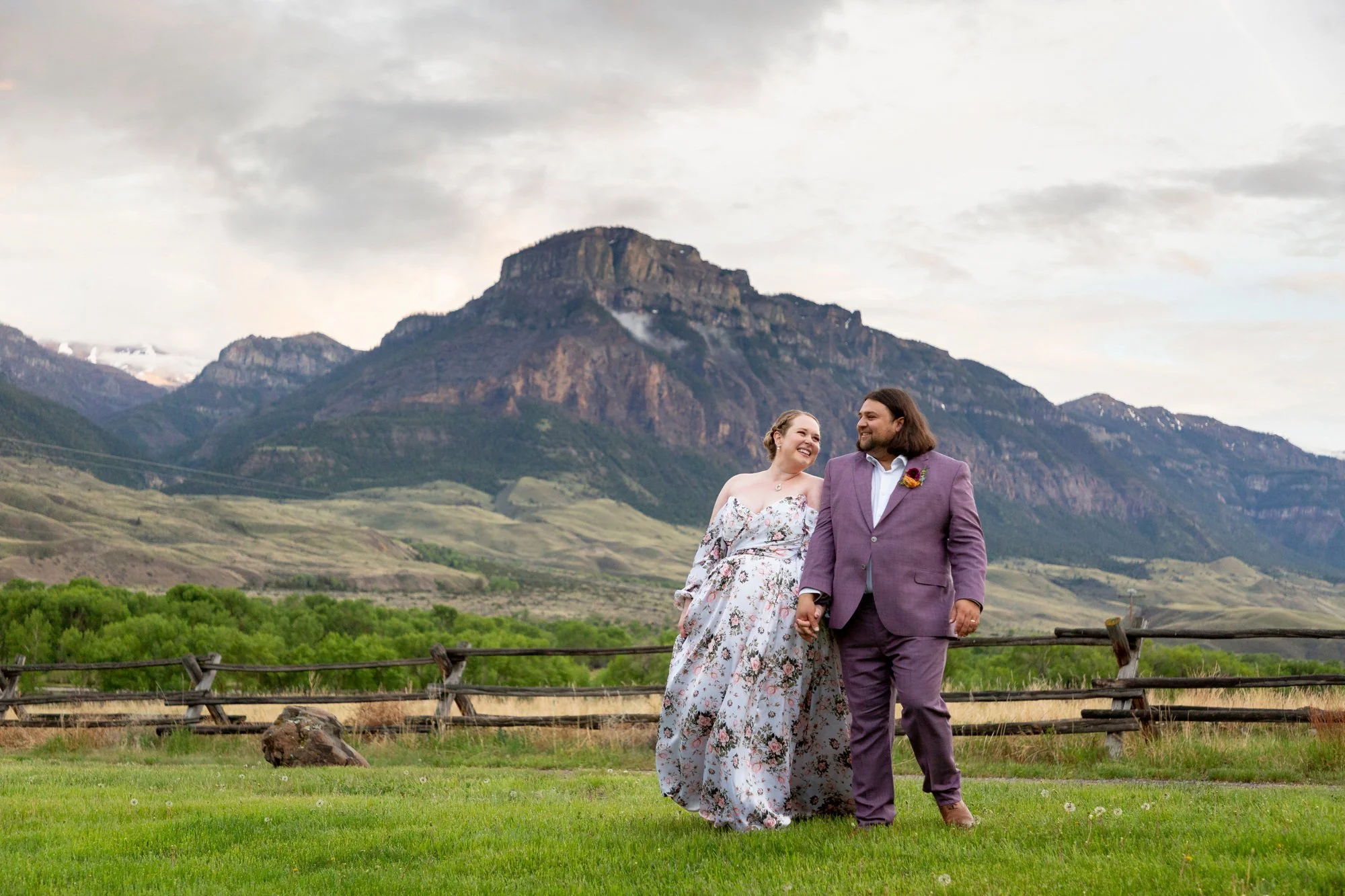 A smiling couple holding hands and walking in a green field with a mountain range in the background, overcast sky.