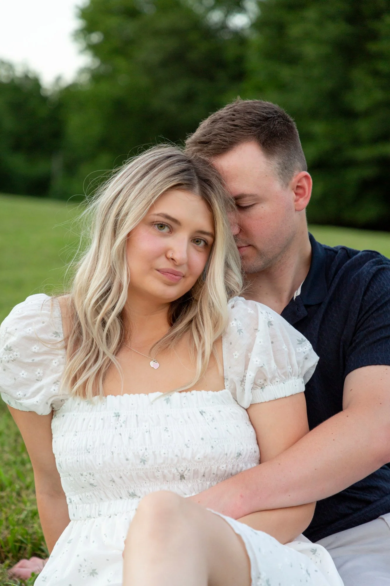 A young couple sitting outdoors on grass with trees in background. The woman has blonde hair, wearing a white dress with floral pattern, and has a nose piercing. The man has short brown hair, wearing a dark shirt, with eyes closed, resting his forehe