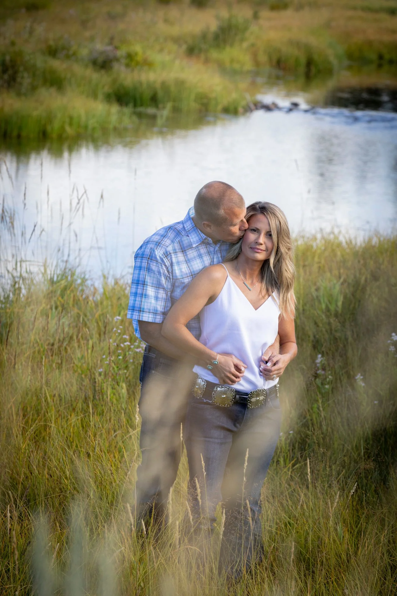 A couple stands in a grassy area near a small lake, with the man kissing the woman on the cheek. The woman is wearing a white sleeveless top and gray pants, while the man is dressed in a blue plaid shirt. The background features water and lush green 