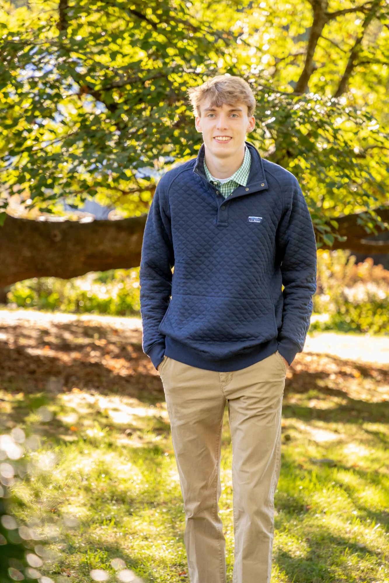 A young man standing outdoors on a sunny day with trees and autumn leaves in the background. He is wearing a navy quilted jacket, a green checkered shirt, and khaki pants, and is smiling at the camera.