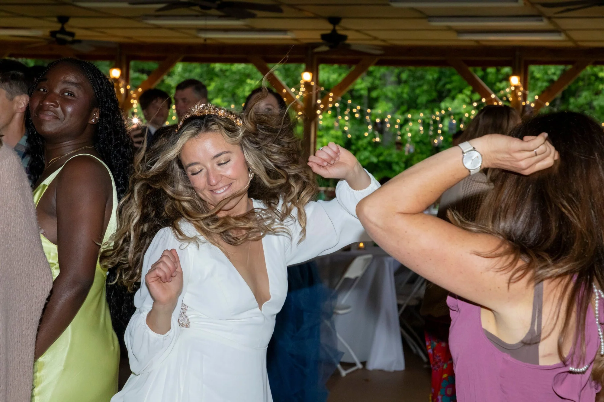 Group of women dancing and enjoying themselves at a festive event under a wooden pavilion decorated with string lights and fans, with greenery visible in the background.