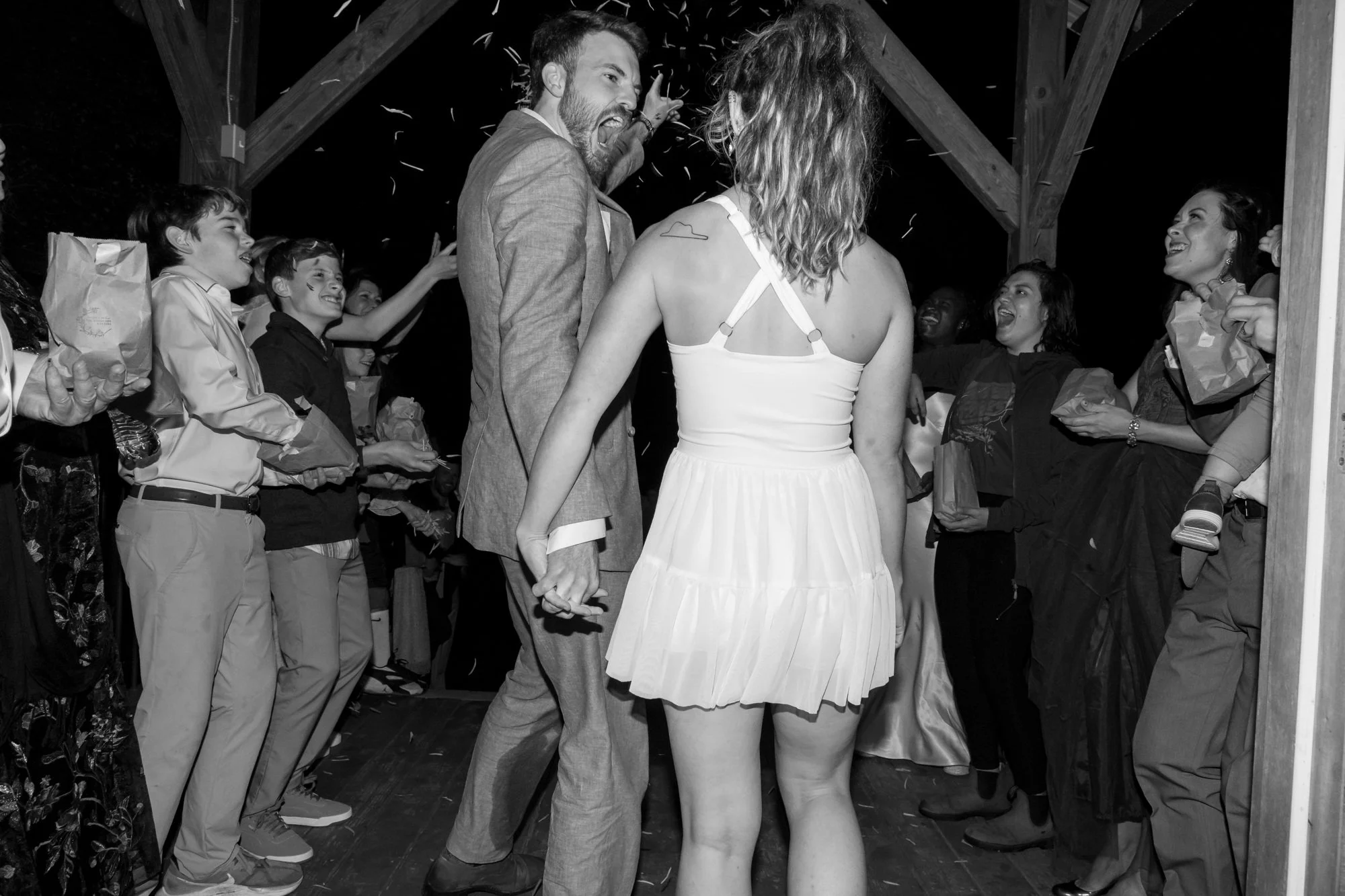 Black and white photo of a couple dancing closely in a wooden interior, surrounded by people joyfully watching and smiling. The woman is wearing a dress with straps, and the man is wearing a blazer.