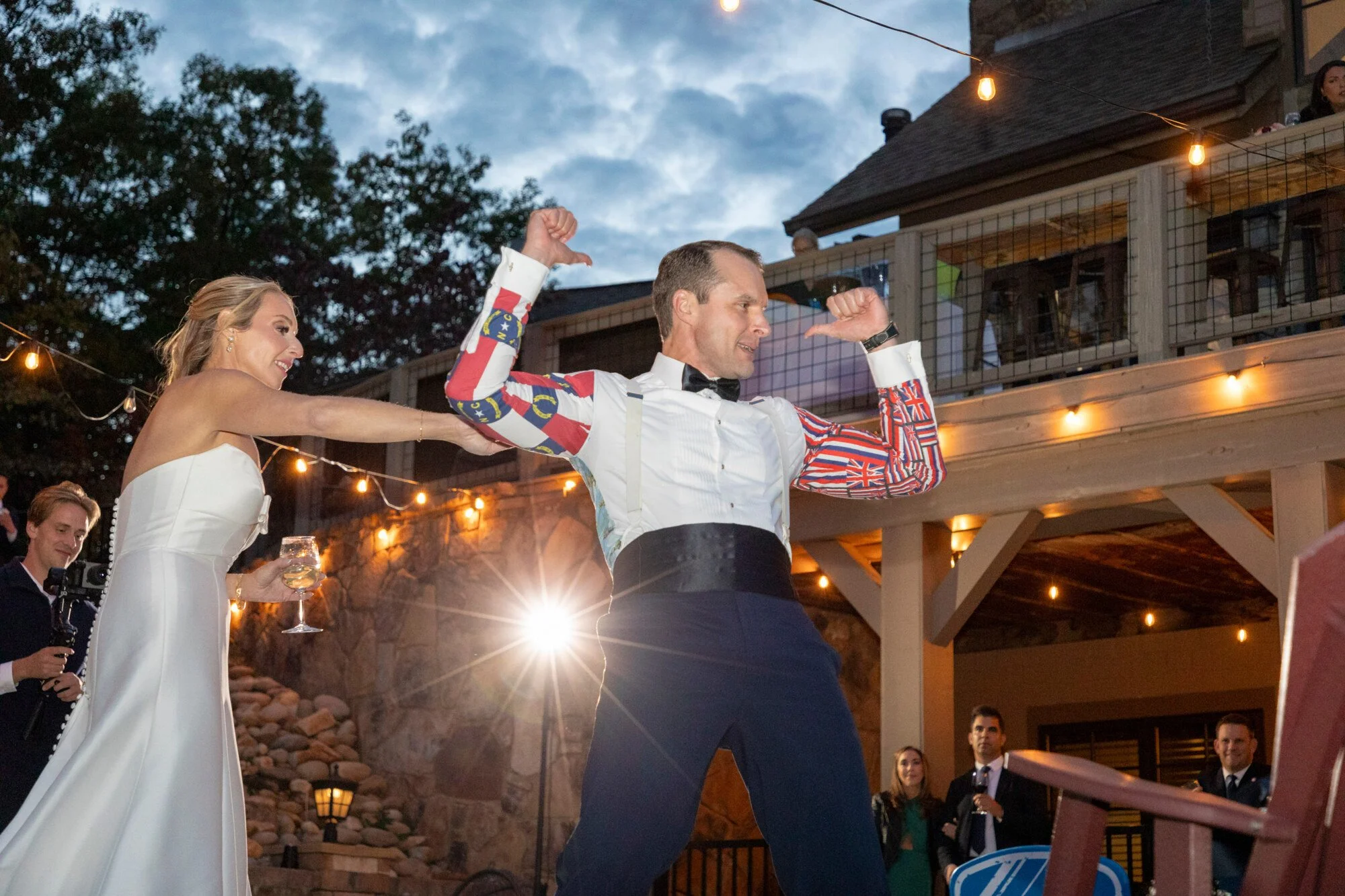 A man and woman dancing at a wedding reception outdoors during evening. The woman is wearing a white wedding dress holding a glass, smiling, while the man is dressed in a tuxedo with an American flag-themed blazer, flexing his muscles. Guests are vis