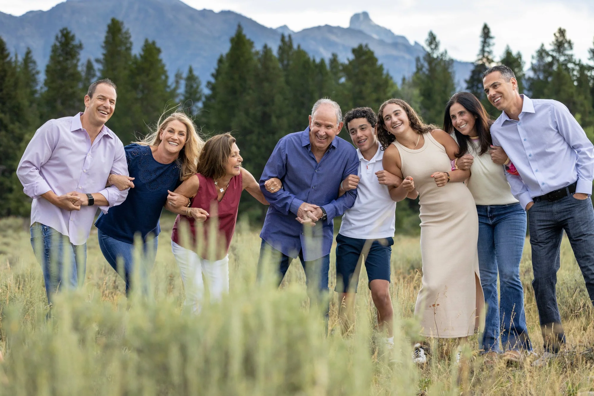 A group of nine people, men and women, standing outdoors in a line, smiling, and holding arms in a scenic outdoor setting with trees and mountains in the background.