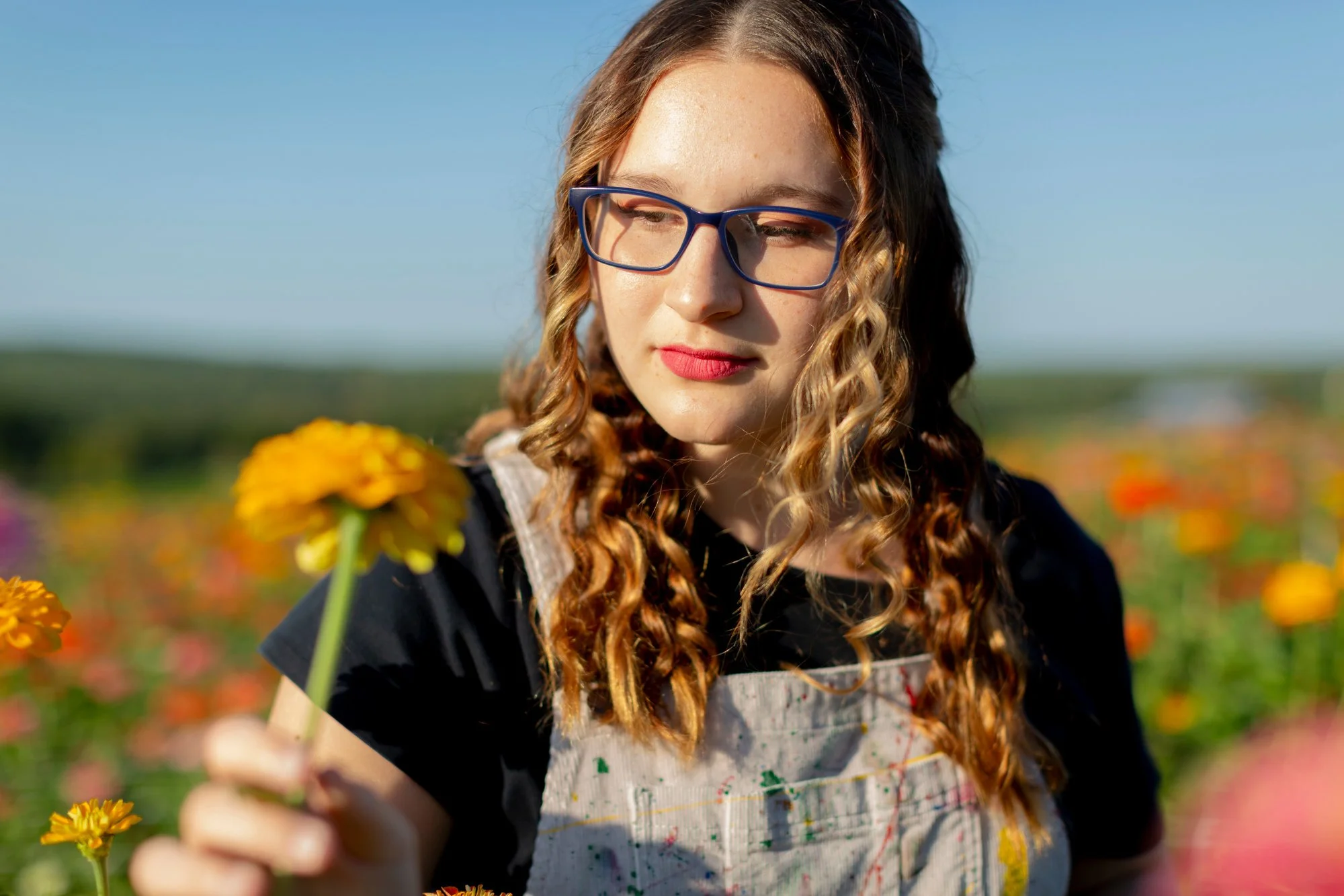 A young woman with curly hair, glasses, and red lipstick in a field of yellow and orange flowers on a sunny day.