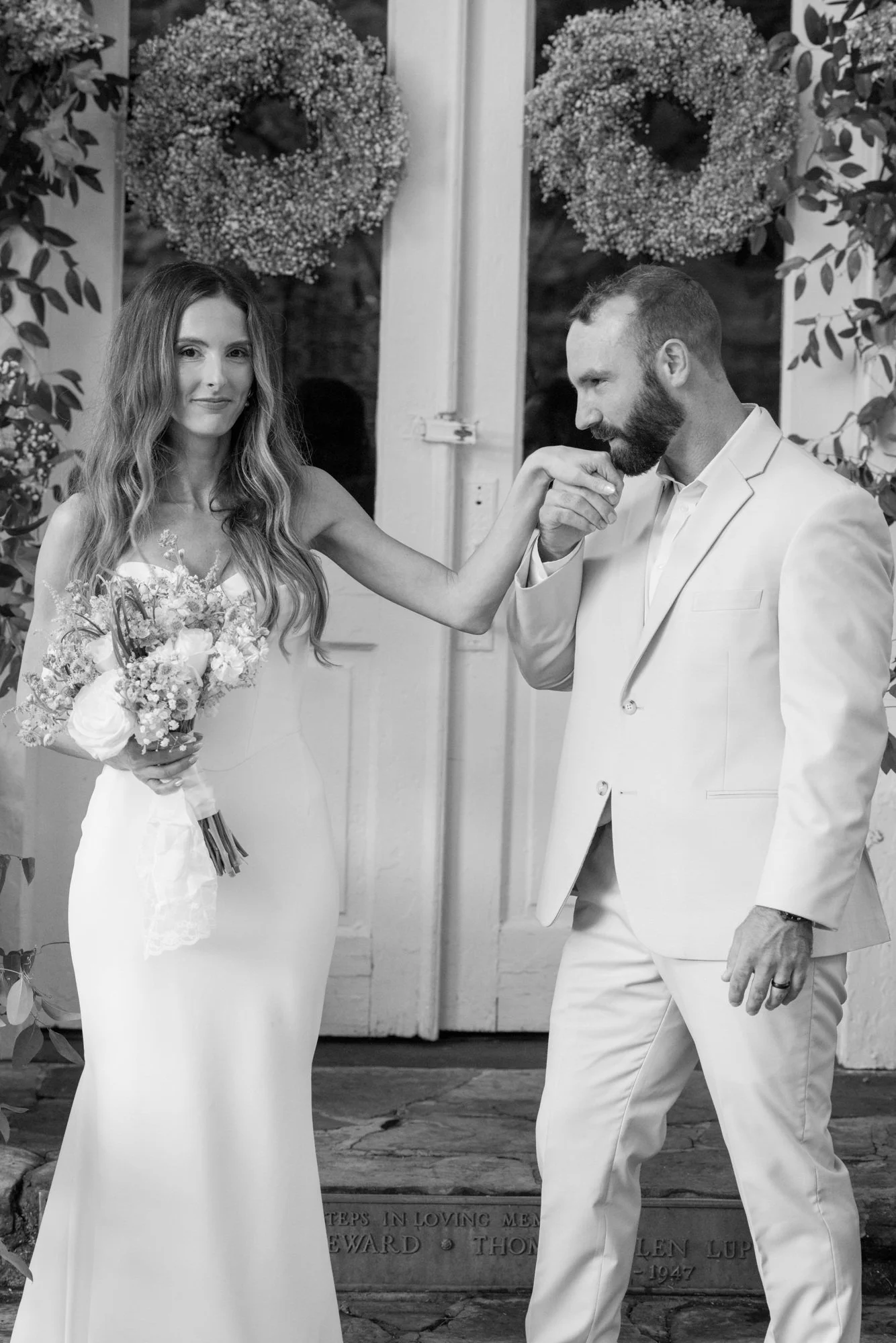 Black and white photo of a bride and groom during their wedding ceremony. The bride has long wavy hair, wears a fitted wedding dress, and holds a bouquet of flowers. The groom has a beard, wears a white suit, and is kissing the bride's hand. They are